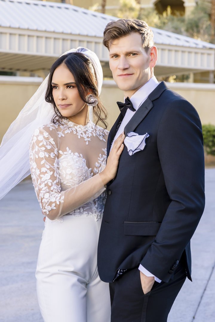 En brud og en brudgom står sammen utendørs, hvor bruden har på seg en hvit bryllupskjole med frynser og en veil, og brudgommen har på seg en svart dress og slips.