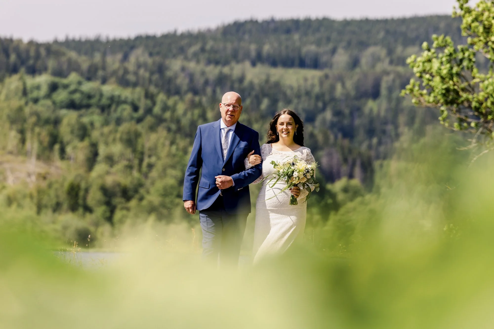 En brud og en eldre mann går utendørs, omgitt av grønt landskap og fjell, på en solrik dag.