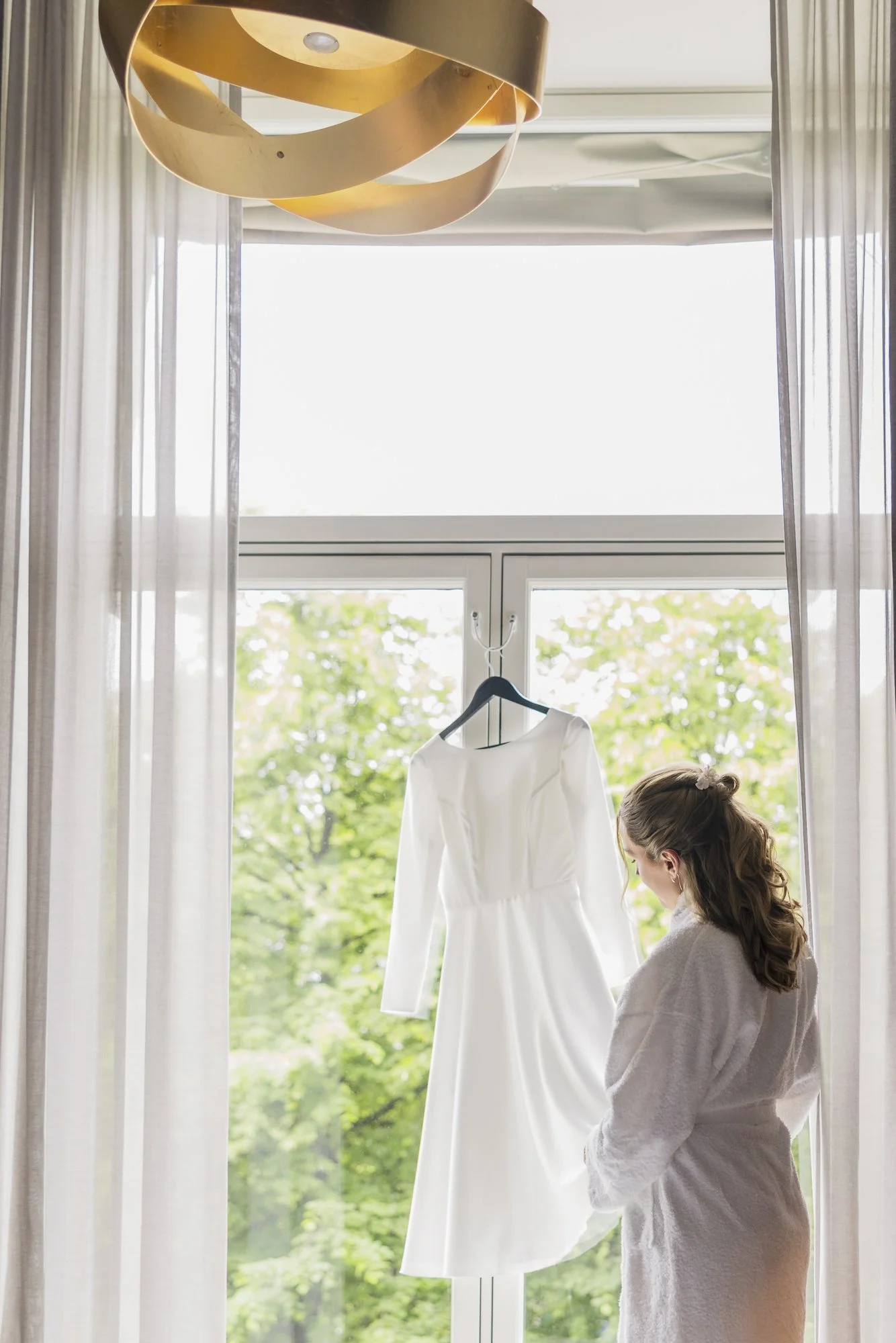 Kvinne i badekåpe ser på en hvit brudekjole hengende foran et vindu med grønne trær utenfor.