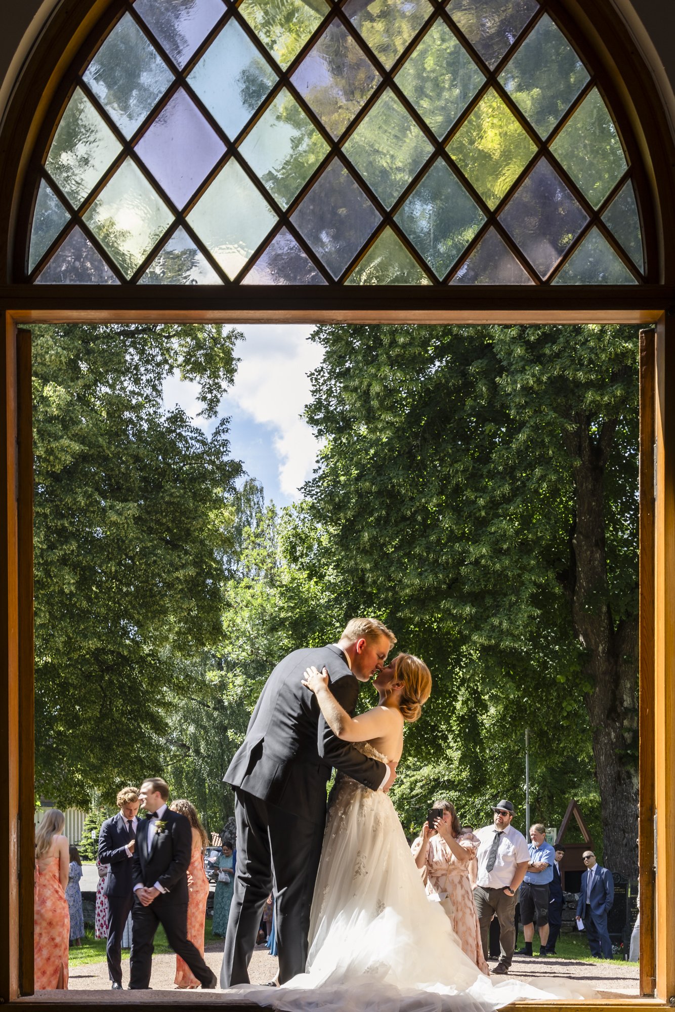 Et brudepar kysser under en stor trebuet arkivglass i en utendørs bryllupsseremoni med gjester i bakgrunnen.