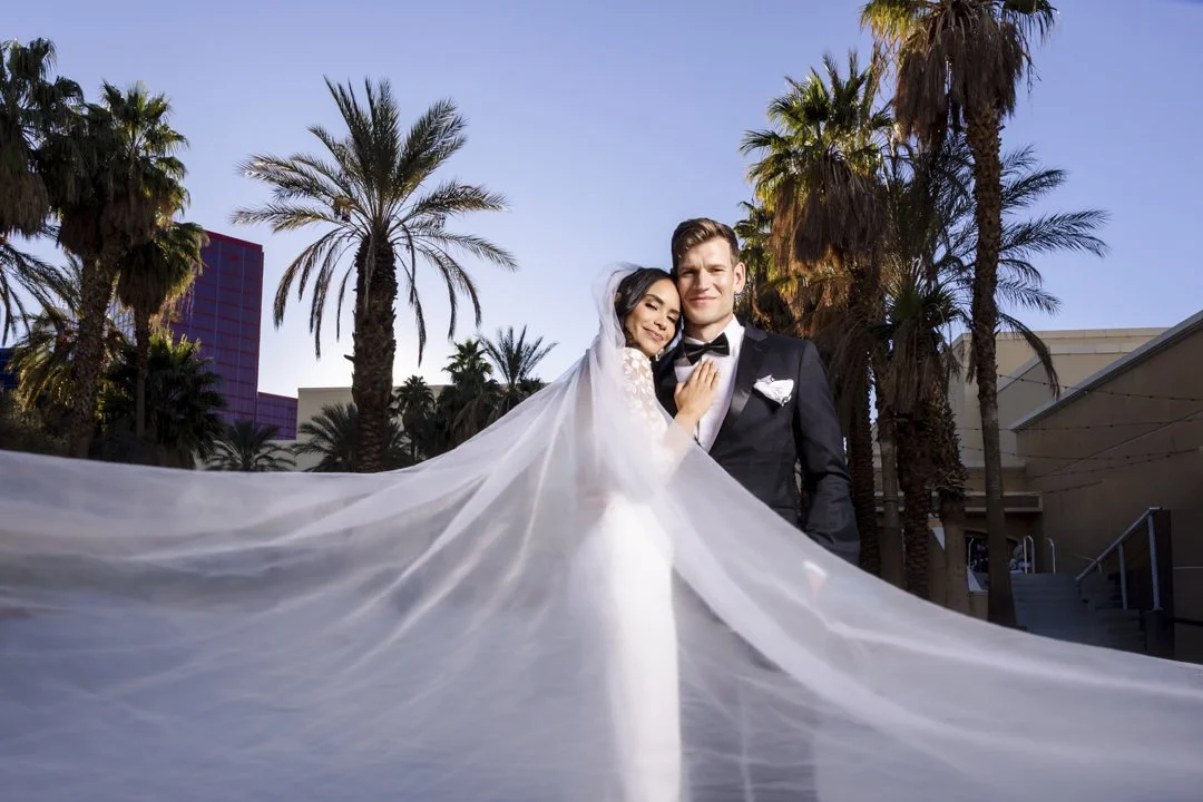 Et nyttgift par i brudeklær står under palmtrær i en utendørs setting.