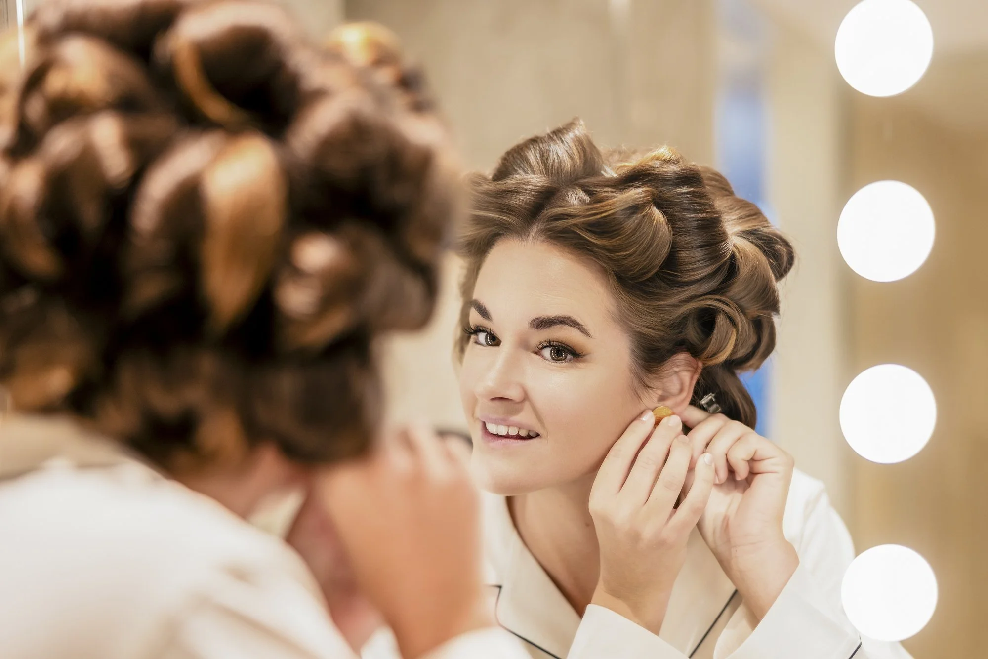 En kvinne sminker seg foran en speil med lys rundt, har på seg en hvit kjele og har krøllete hår.