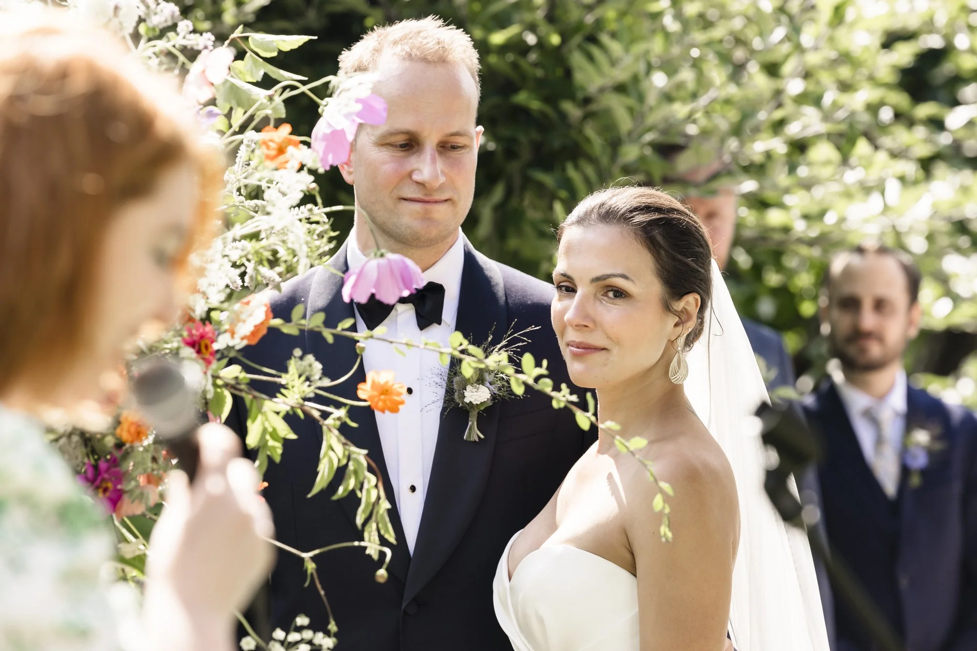 Et bryllup seremonimiljø med bruden og brudgommen som står sammen, omringet av blomster og venner.