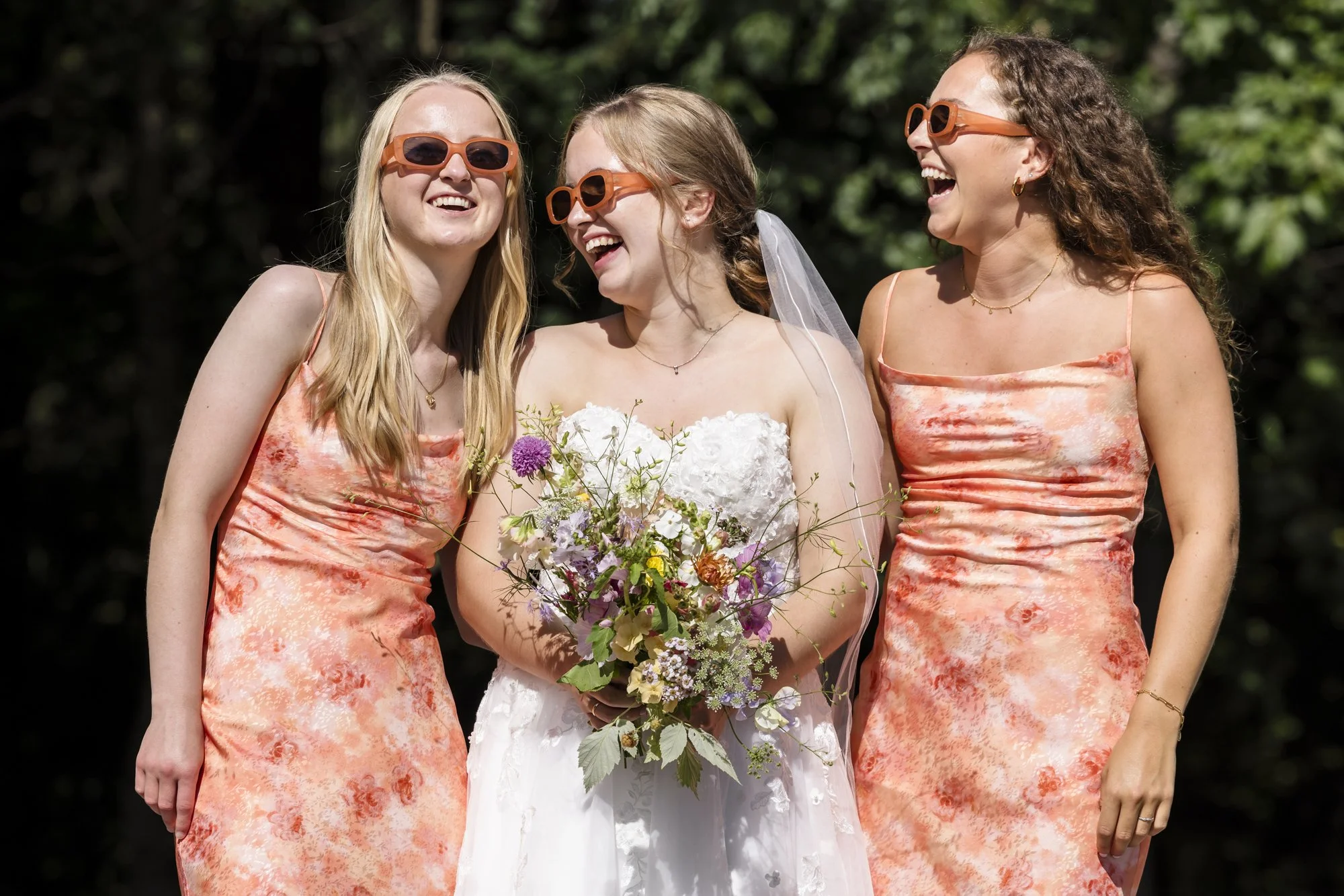 Tre kvinner i brudepikekjoler og brudekjole ler og holder en blomsterbukett utendørs.