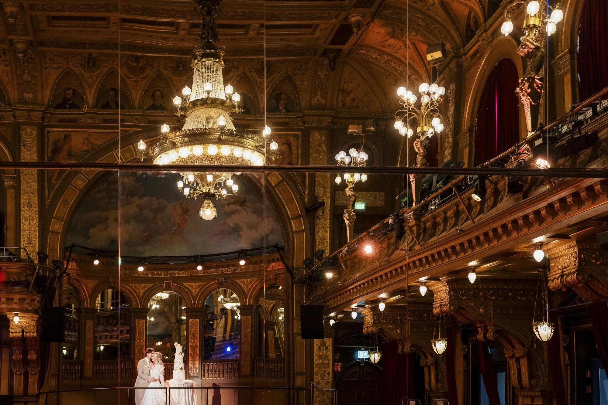Et brudepar står sammen foran en bryllupskake i en overdådig, klassisk sal med store lysekroner, utsmykkede gulldekorasjoner og speilvegger.