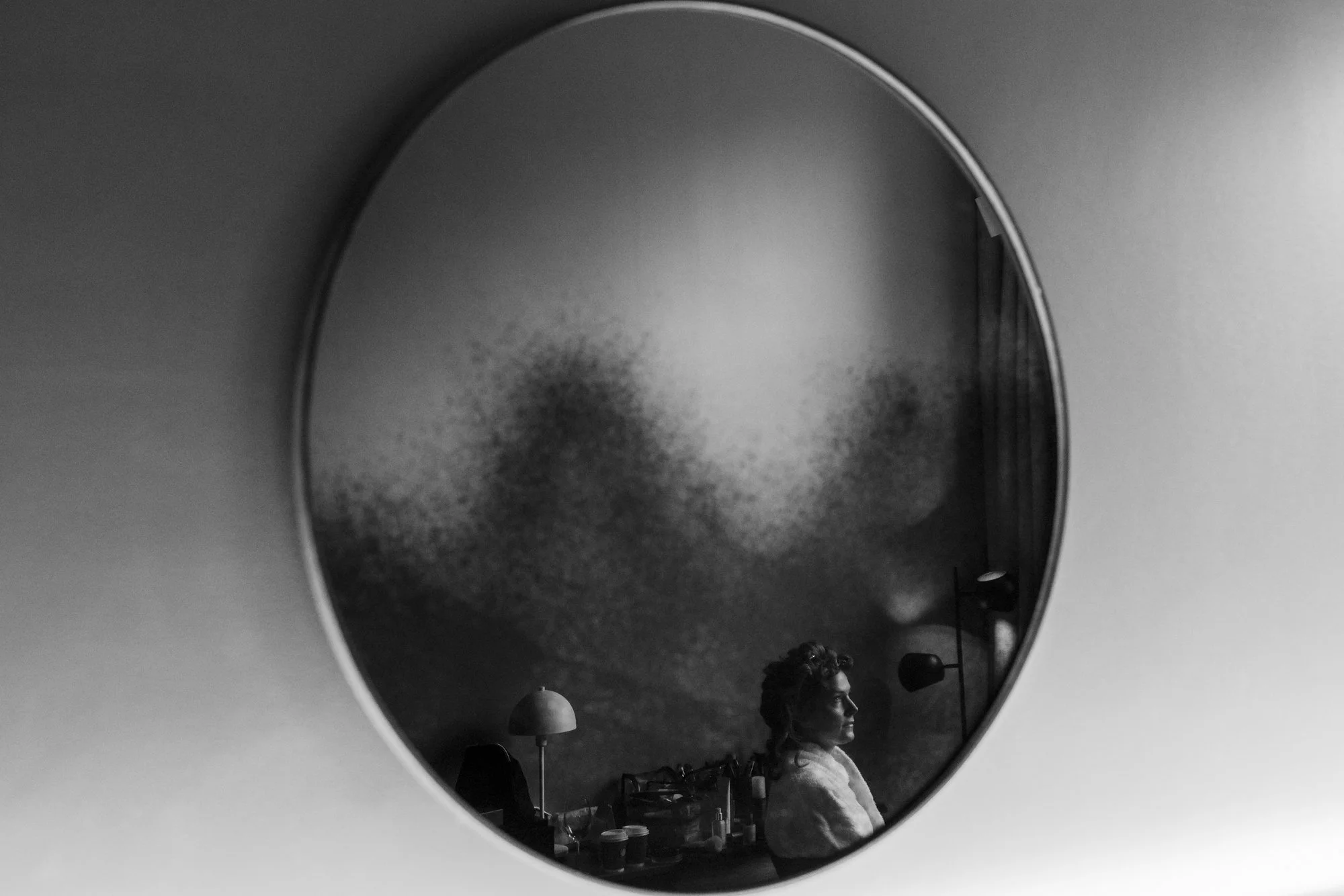 En person ser på seg selv i et rundt speil, med lamper og ulike gjenstander på en hodeskaps overflate i rommet.