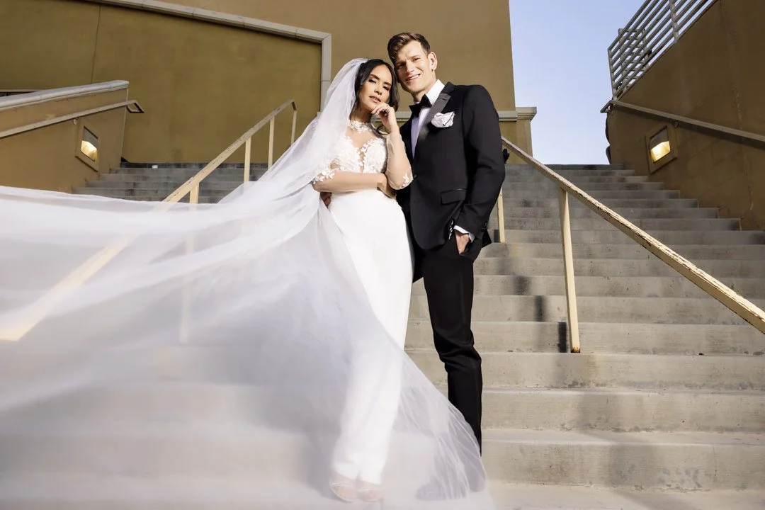 Et brudepar står på trappene utendørs, med kvinnen i en hvit bryllupskjole og mannen i en svart smoking, solnedgang i bakgrunnen.