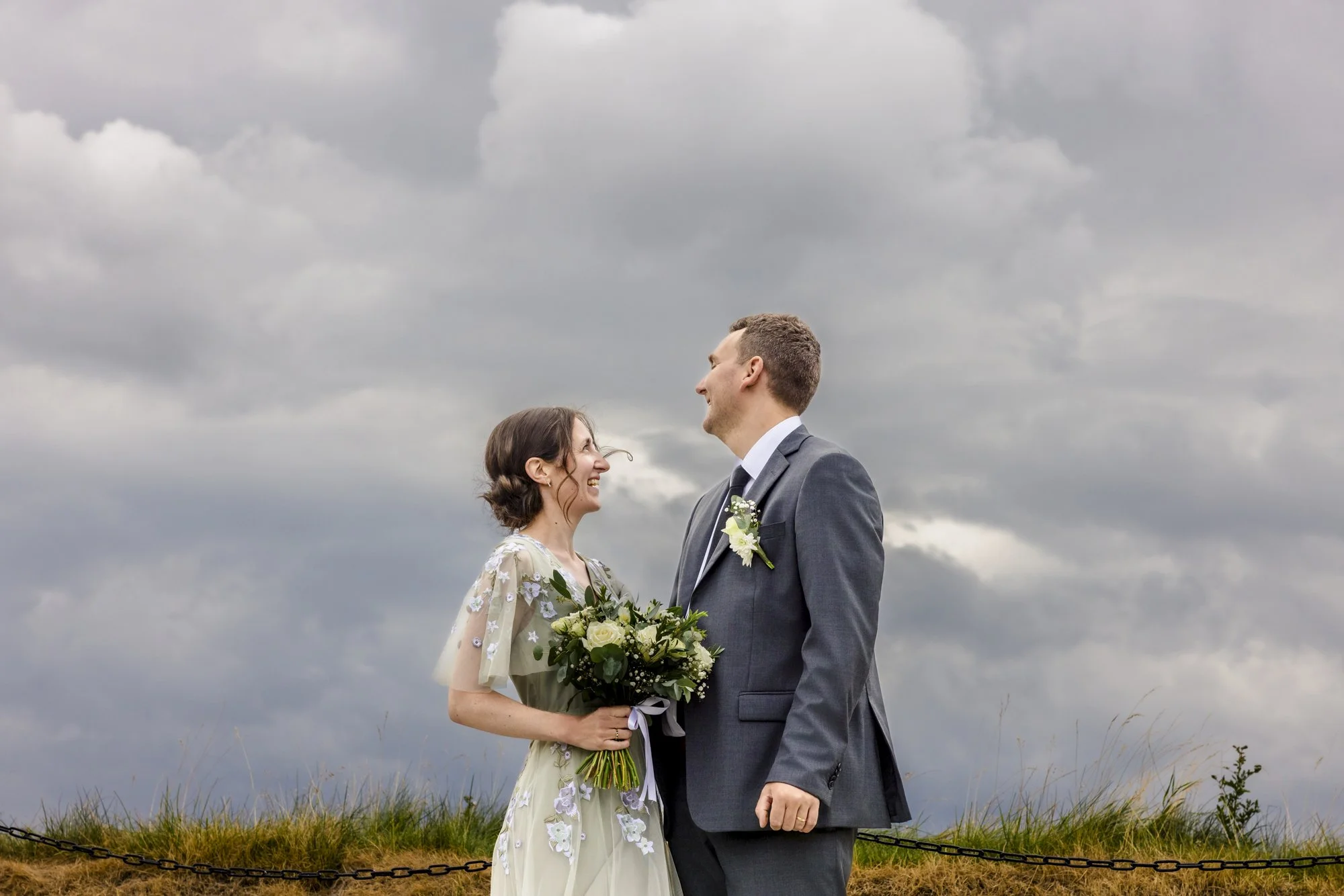 Et par står på en åpen gresslette under en overskyet himmel, kledd i formelle klær og smiler mot hverandre, tydelig forelsket.