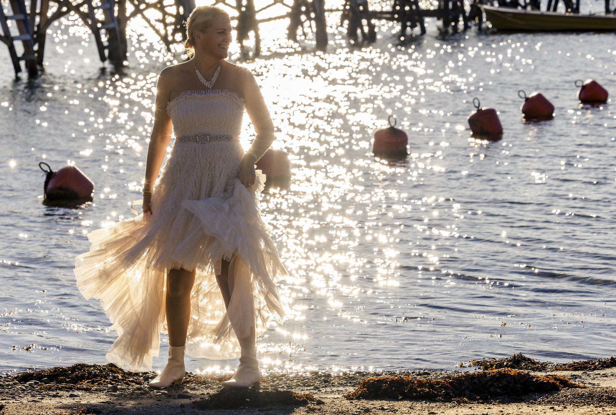 En kvinne i bryllupskjole står på en strand ved vannet, med sollys som reflekteres i vannet og båt og bøyer i bakgrunnen.