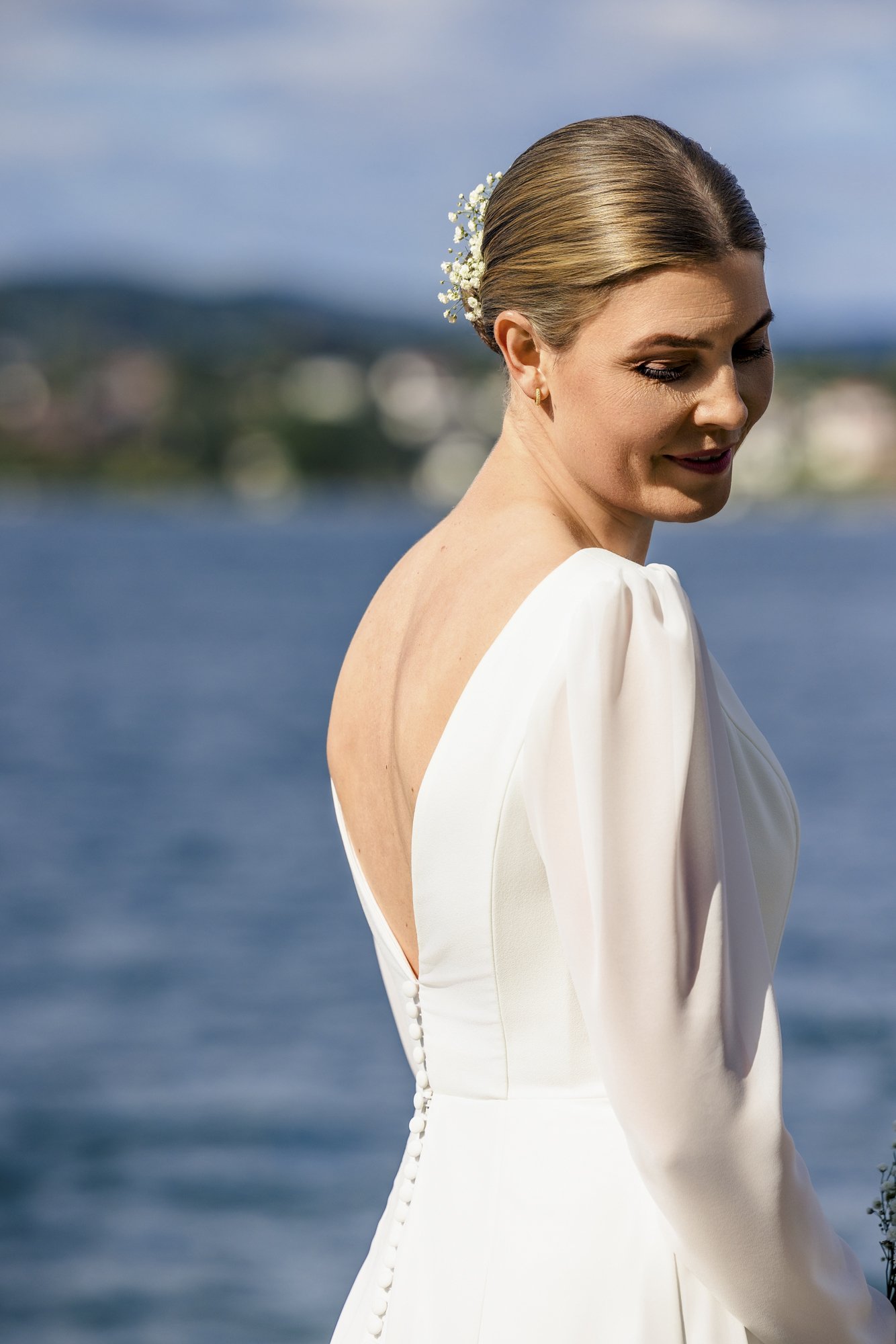 Nydelig kvinne i hvit brudekjole med oppsatt hår pyntet med hvite blomster, står ved vann på en klar dag.