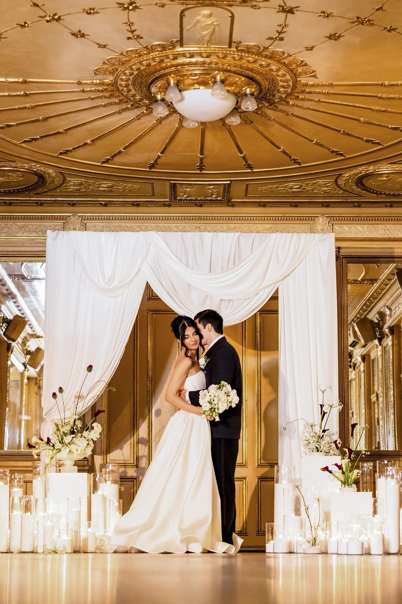 Et nytt ektepar står sammen under en pyntet ramme med hvide gardiner, omgitt av lysende lyslykter og blomster, i en elegant, gild rosesal.