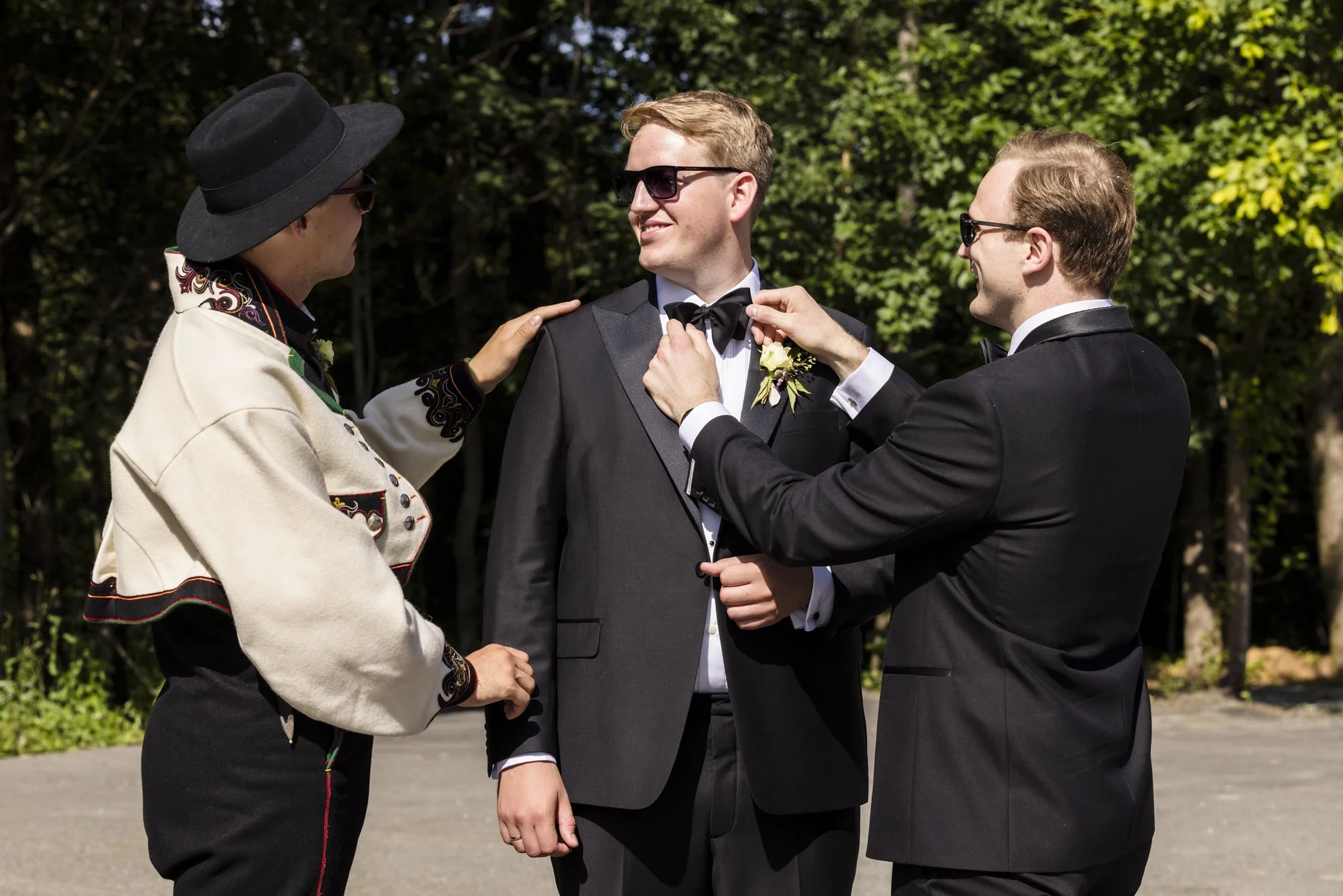 Tre menn i formell dress og solbriller som ler og snakker, ett av dem hjelper en annen med å justere en svart sløyfe. De står utendørs med trær i bakgrunnen.