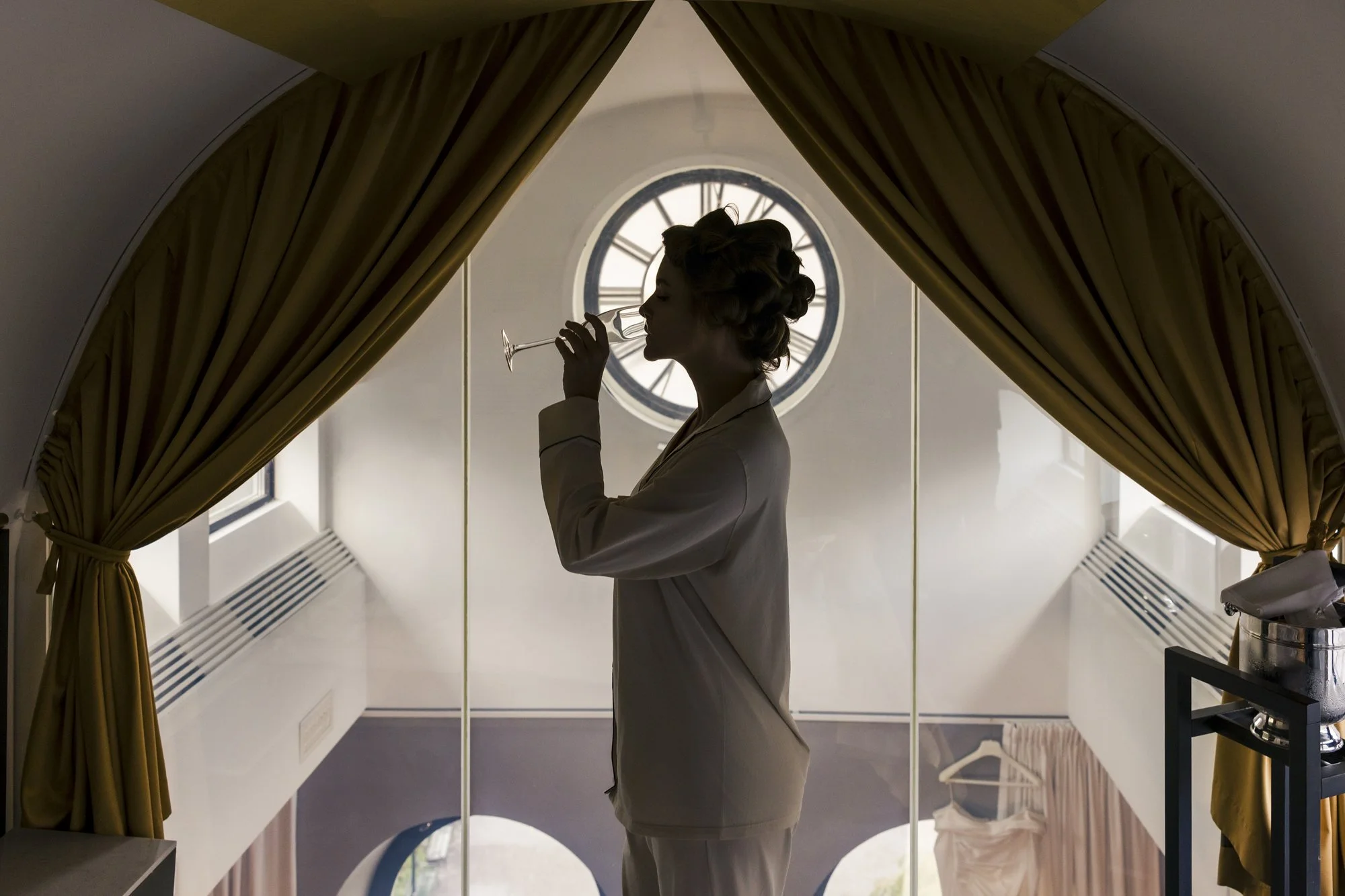 Silhuett av en kvinne som drikker fra et champagneglass foran et stort klokke-vinge, sett fra innsiden av et rom med gule gardiner.