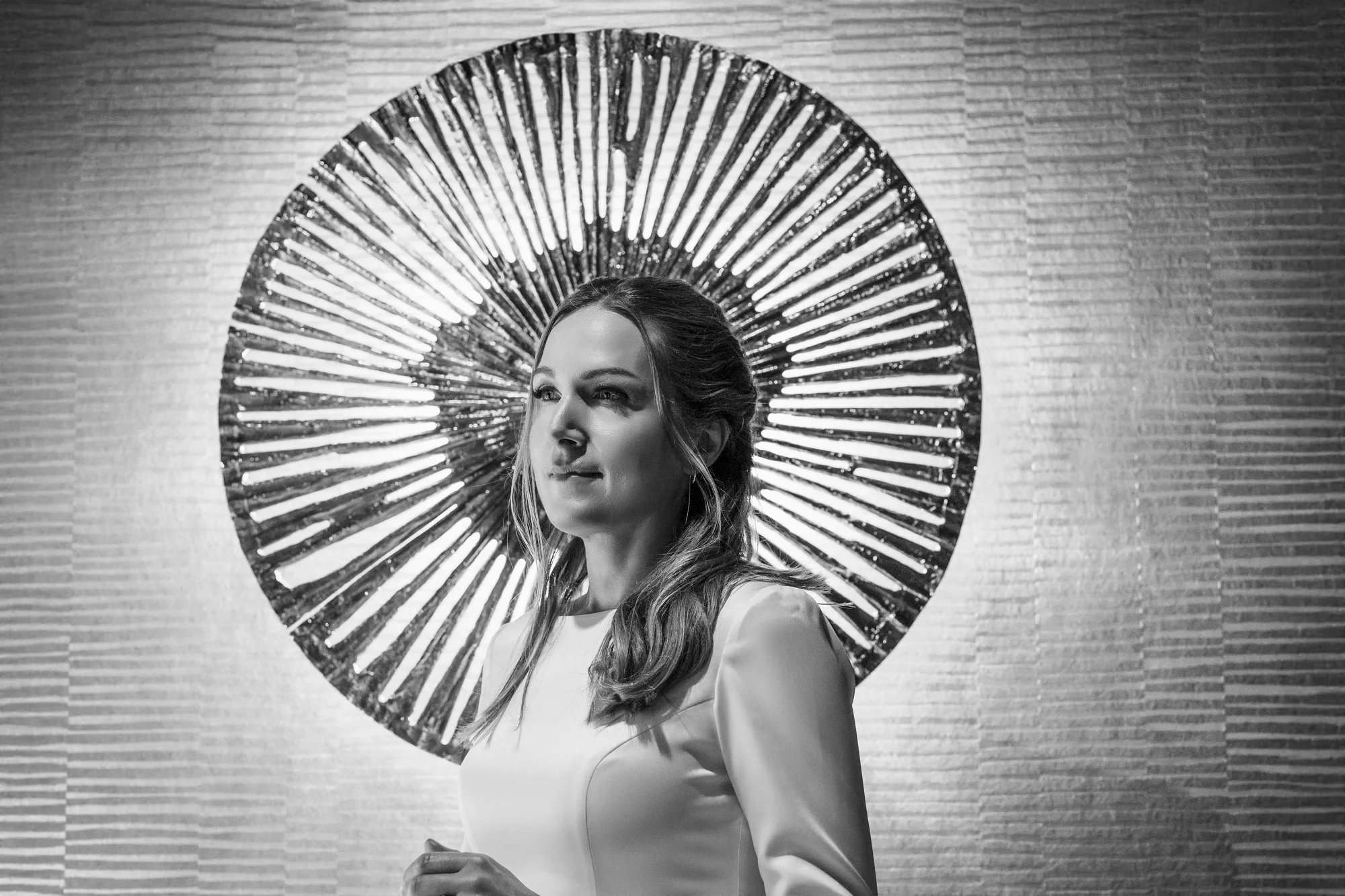 Sort-hvitt fotografi av en kvinne med langt hår, stående foran en dekorativ vegg og en stor, sirkulær metallskulptur bak hodet hennes.