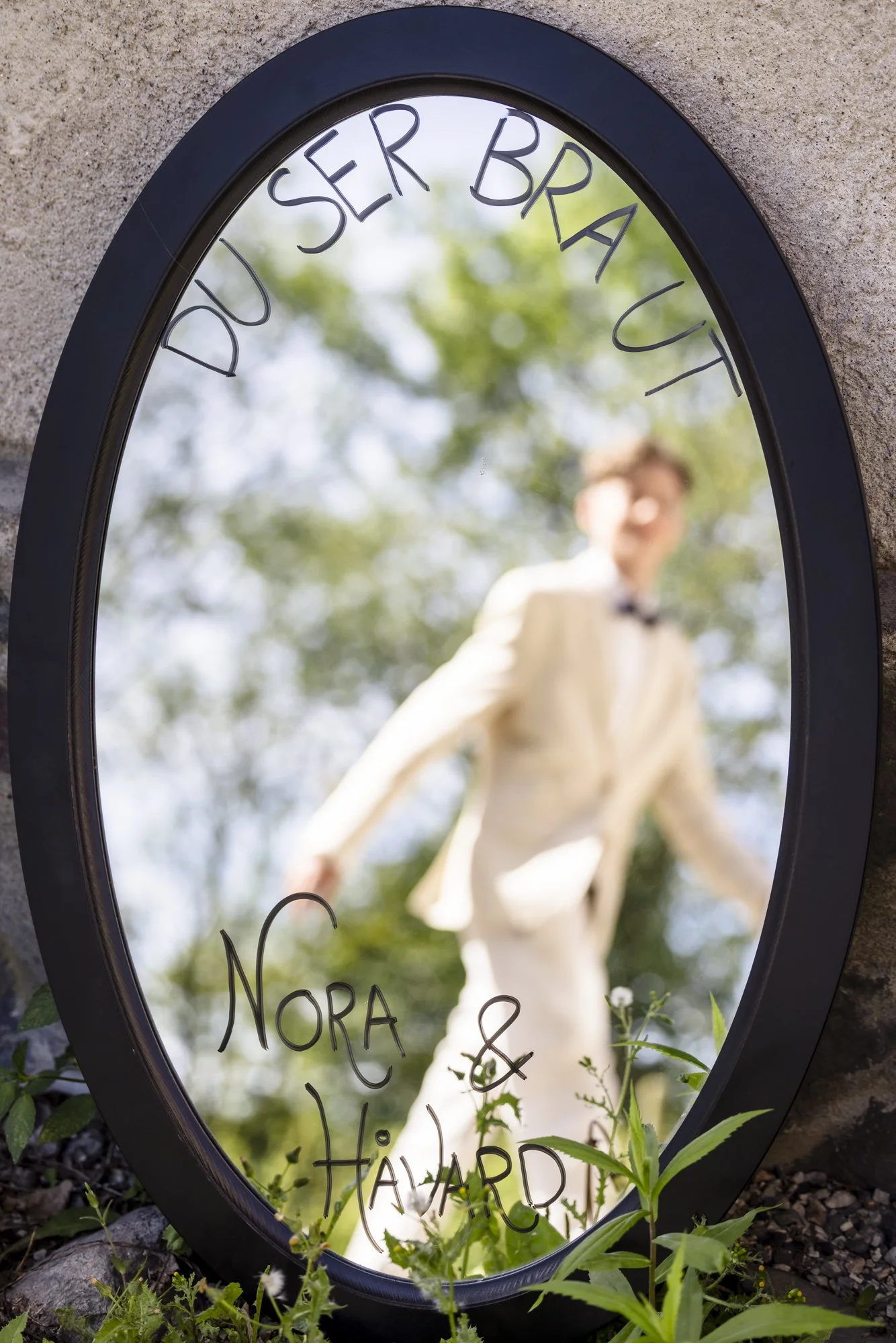 Refleksjon i en oval speil med teksten "DU SER BRA UT" og "NORA & HÅvard", skue ind i en person i en lys dress, omgitt av grønt planteliv.