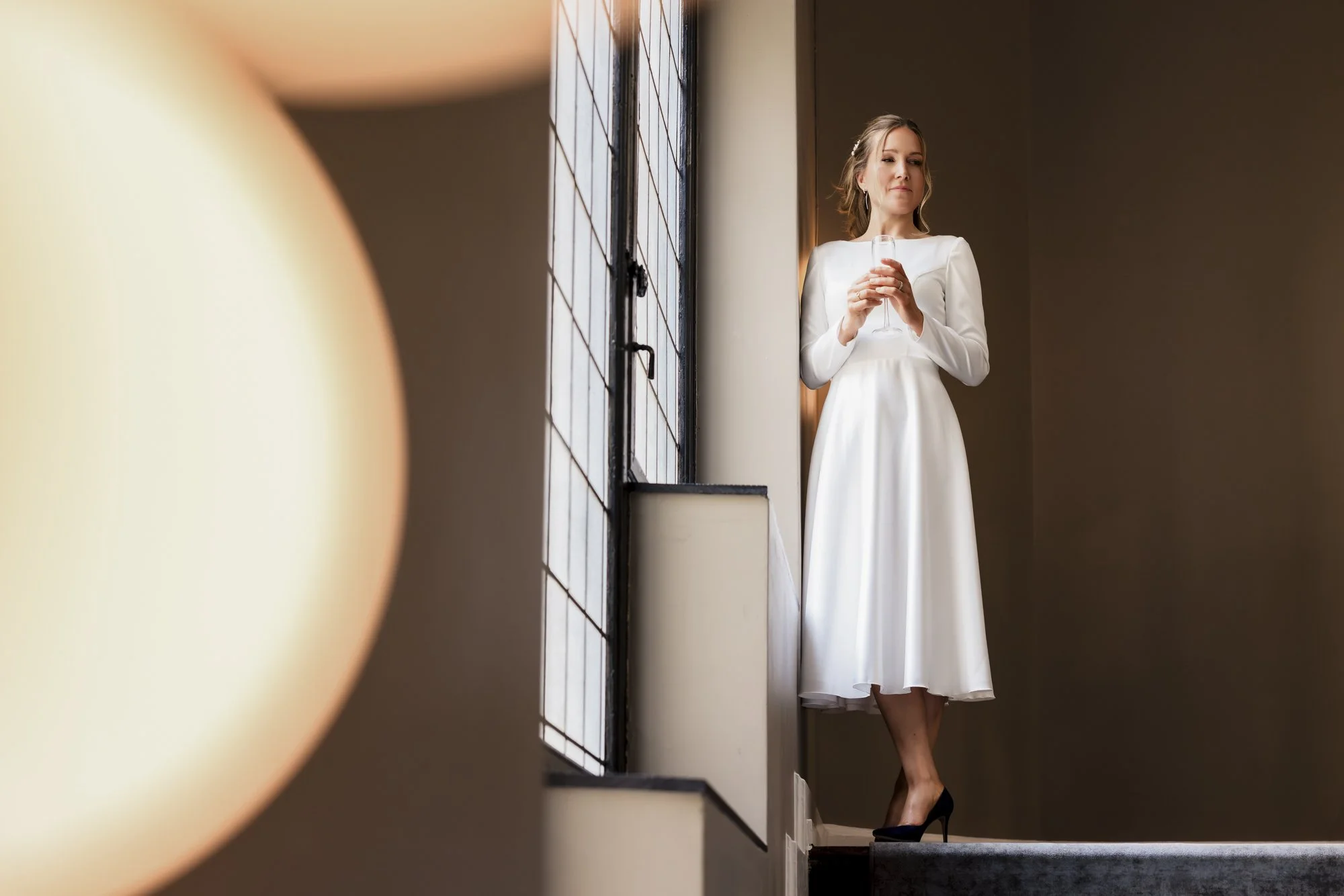 En kvinne i hvit kjole og sorte høyhælte sko står ved et vindu og holder et glass, ser ut til å vente eller nyte en drink.