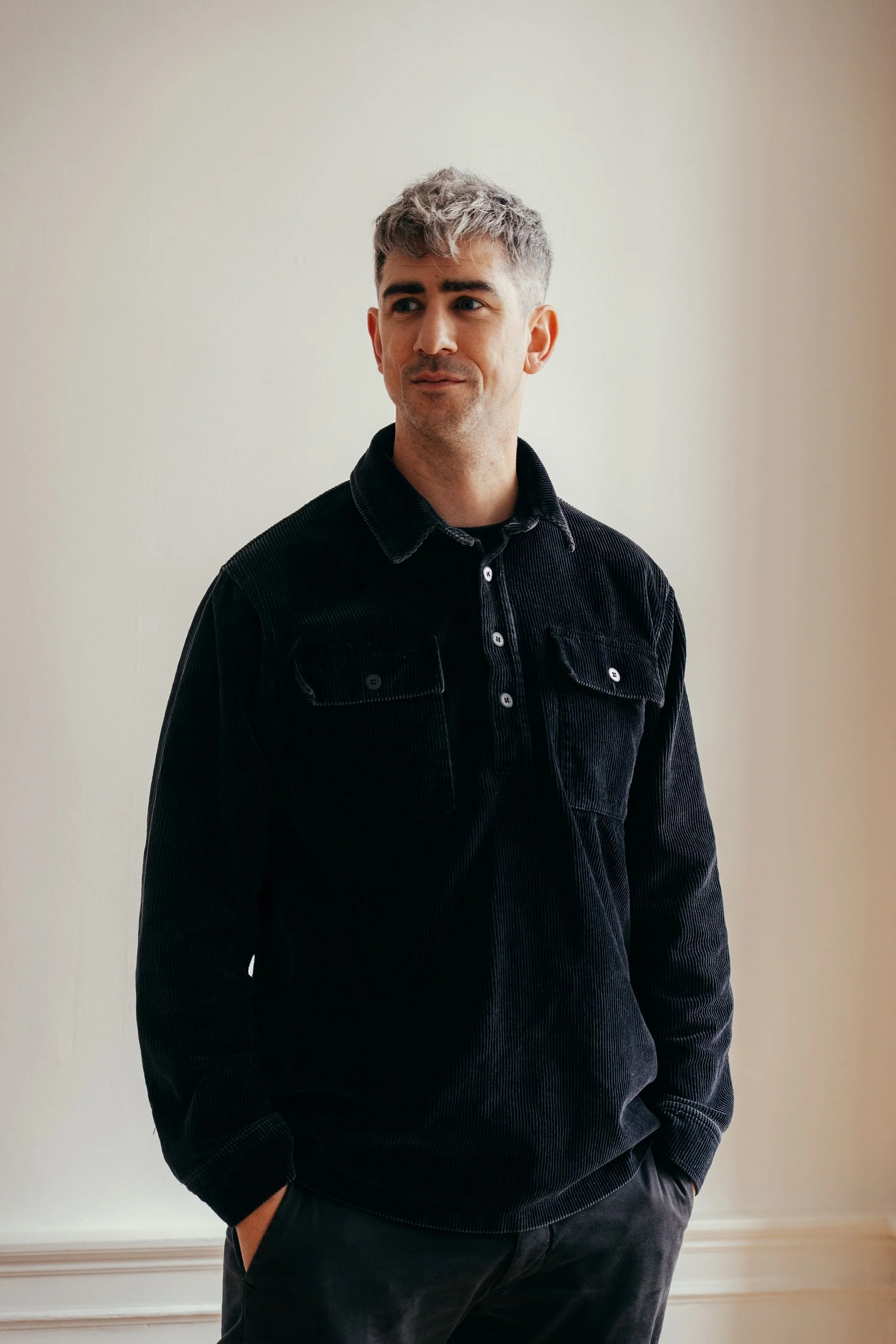 A man with short, gray hair and blue eyes wearing a black corduroy shirt and black pants, standing indoors against a plain off-white wall.