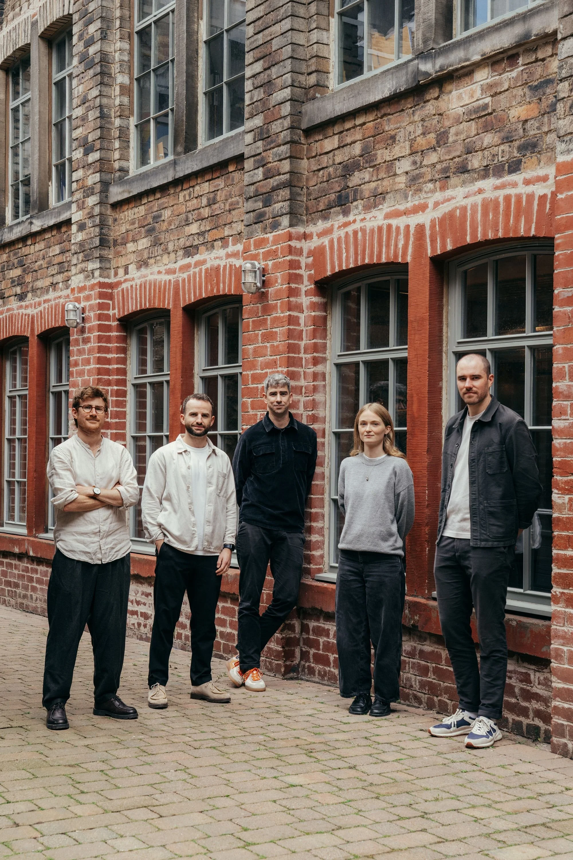 Five young adults standing outdoors in front of a red brick building with large windows, posing for a group photo.