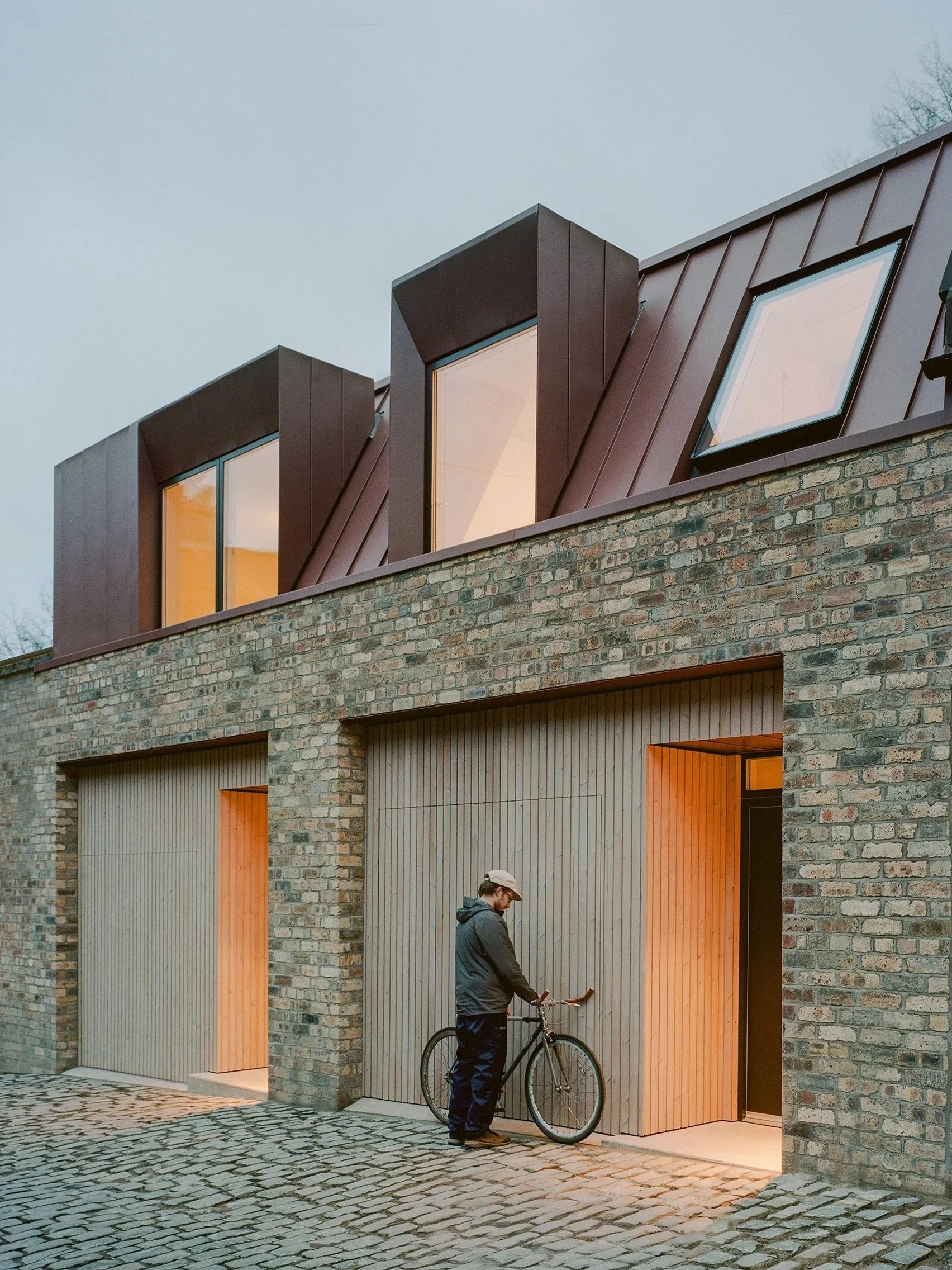 A man in a gray jacket and black pants, wearing a beige cap, stands with his bicycle outside a modern building with brick walls and wood-paneled garage doors, under a cloudy sky.