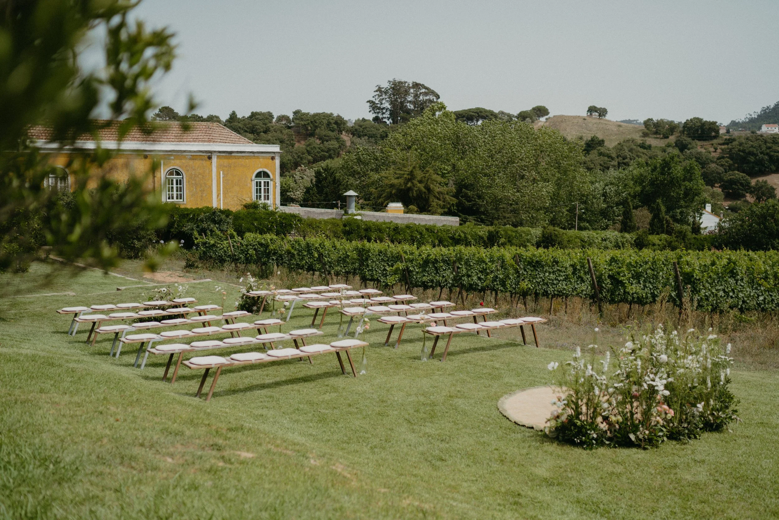 Outdoor wedding ceremony setup with wooden benches facing a circular floral arrangement, surrounded by greenery and a yellow building.