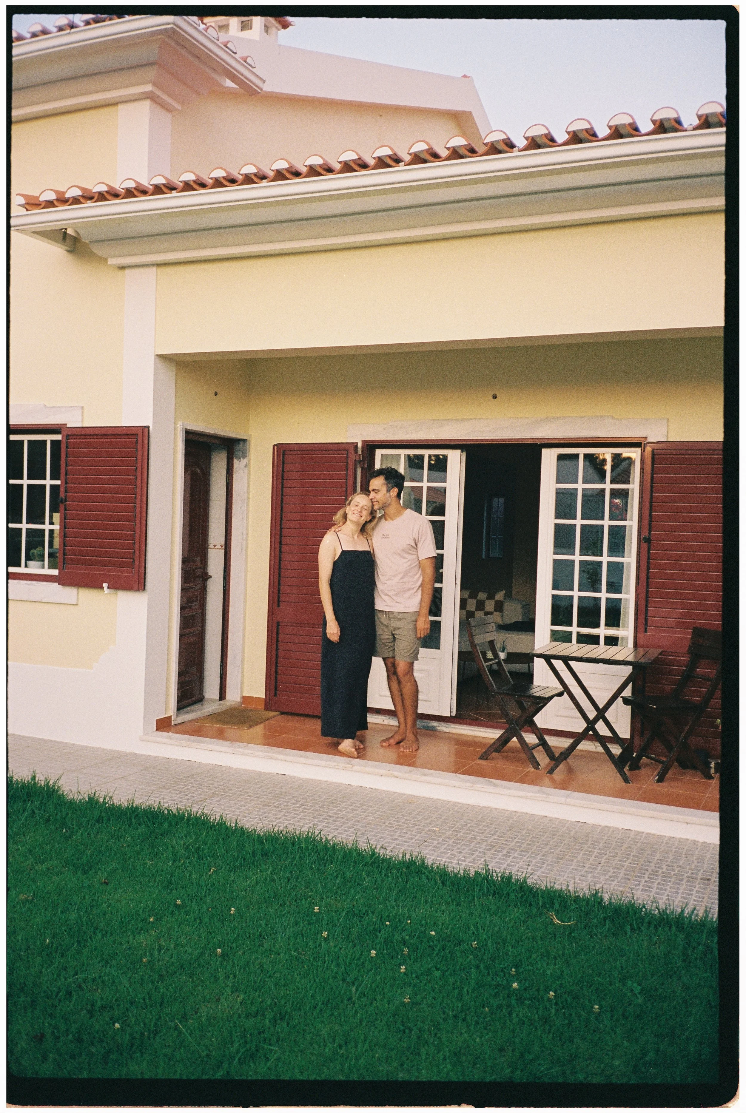 A couple standing on a porch outside a house, smiling and leaning against each other with closed eyes.