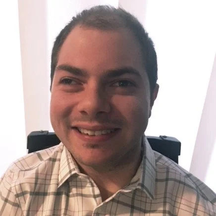 A man smiling, wearing a plaid shirt, seated indoors.