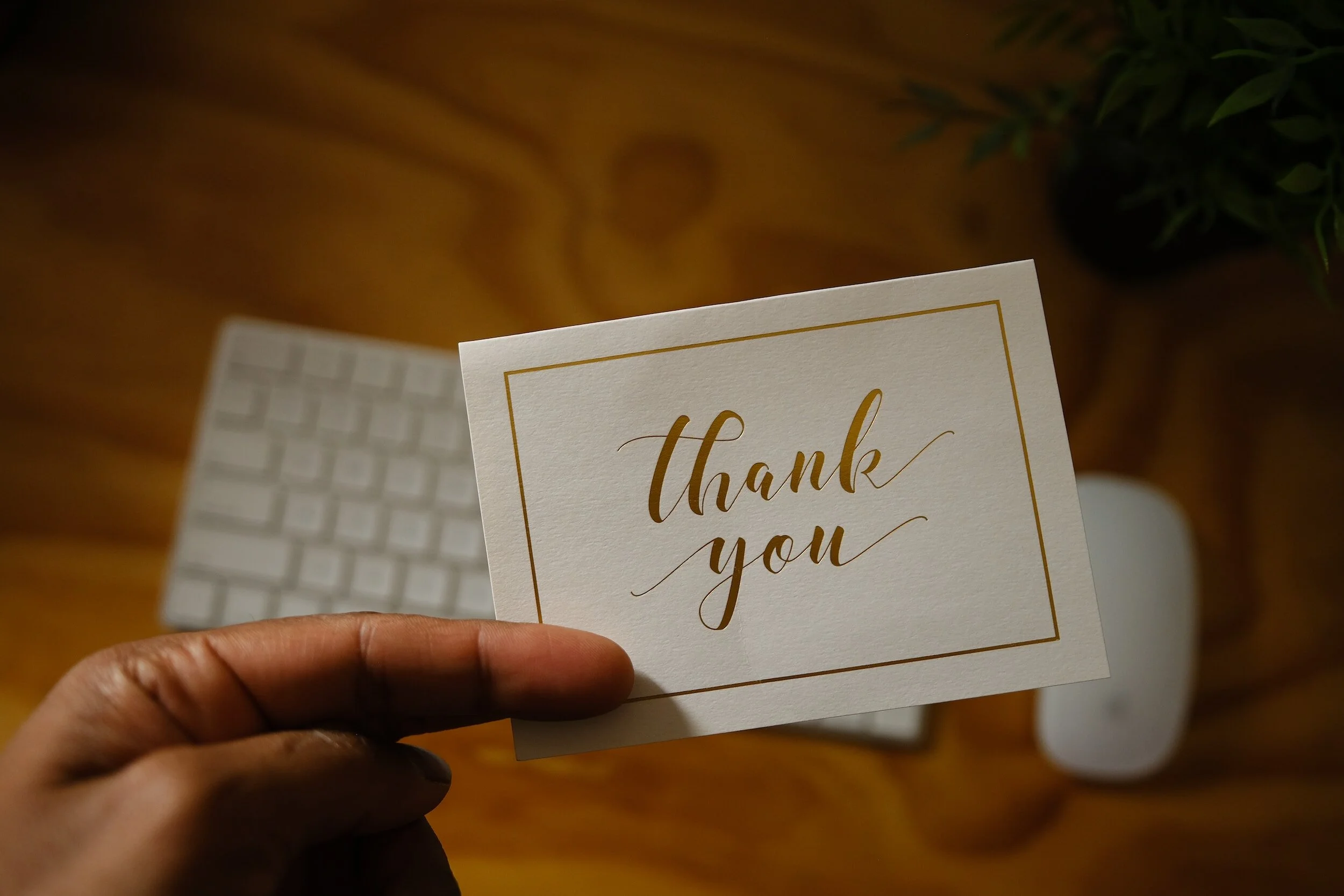 A hand holding a white thank you card with gold lettering, against a blurred background of a keyboard and a potted plant.