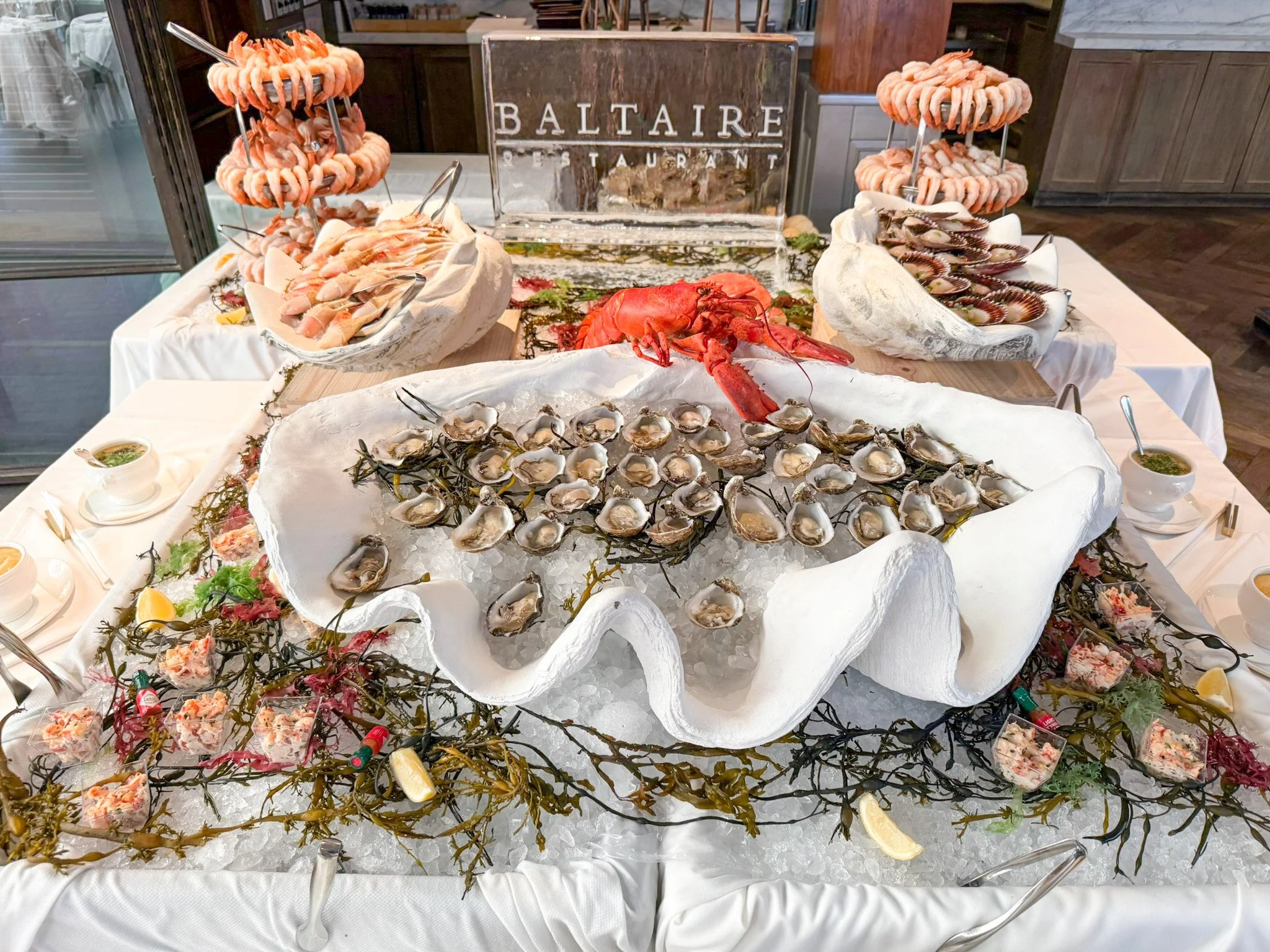 A photo of a grand raw seafood table with shucked oysters on the halfshell inside a giant clam, a lobster, iced sculpture with Baltaire's logo, towers of shrimp, scallops and crab claws