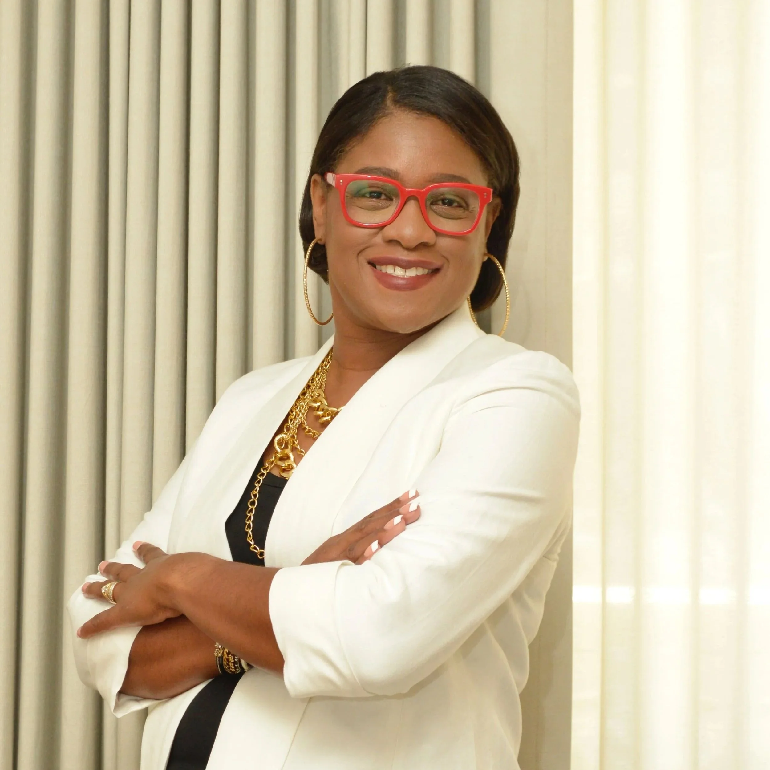 A confident woman with short black hair wearing red glasses, gold hoop earrings, a gold chain necklace, a white blazer, and a black top, smiling with arms crossed in front of cream-colored curtains.