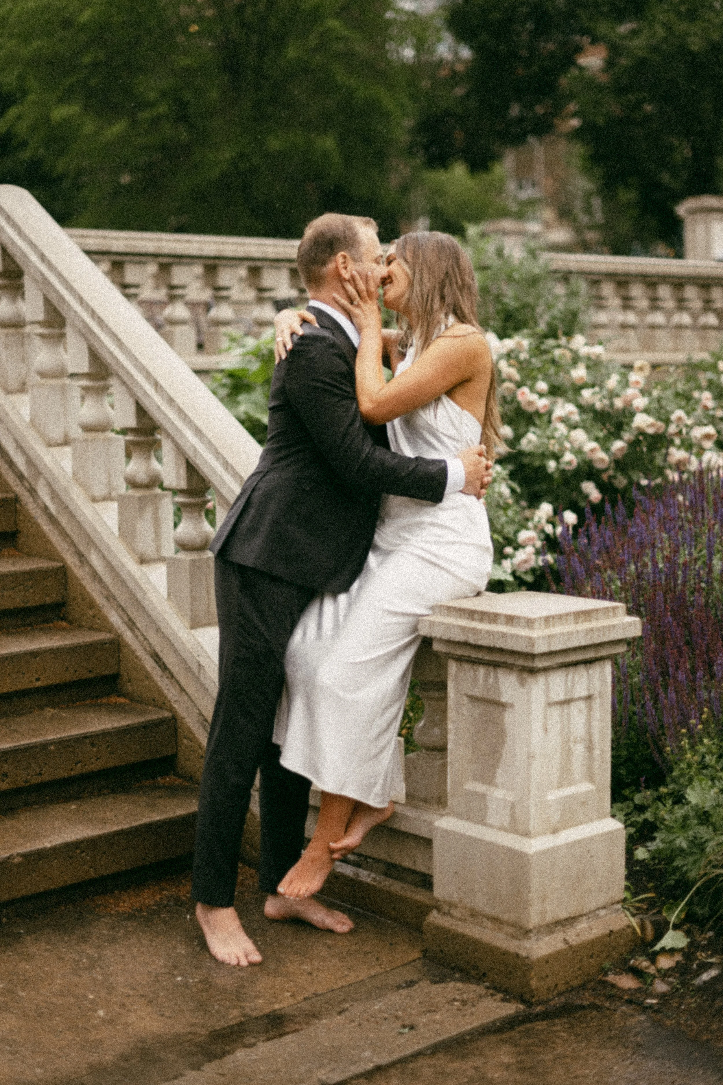 A couple dressed in wedding attire sharing a kiss on a stairway outdoors with flowers and greenery in the background.