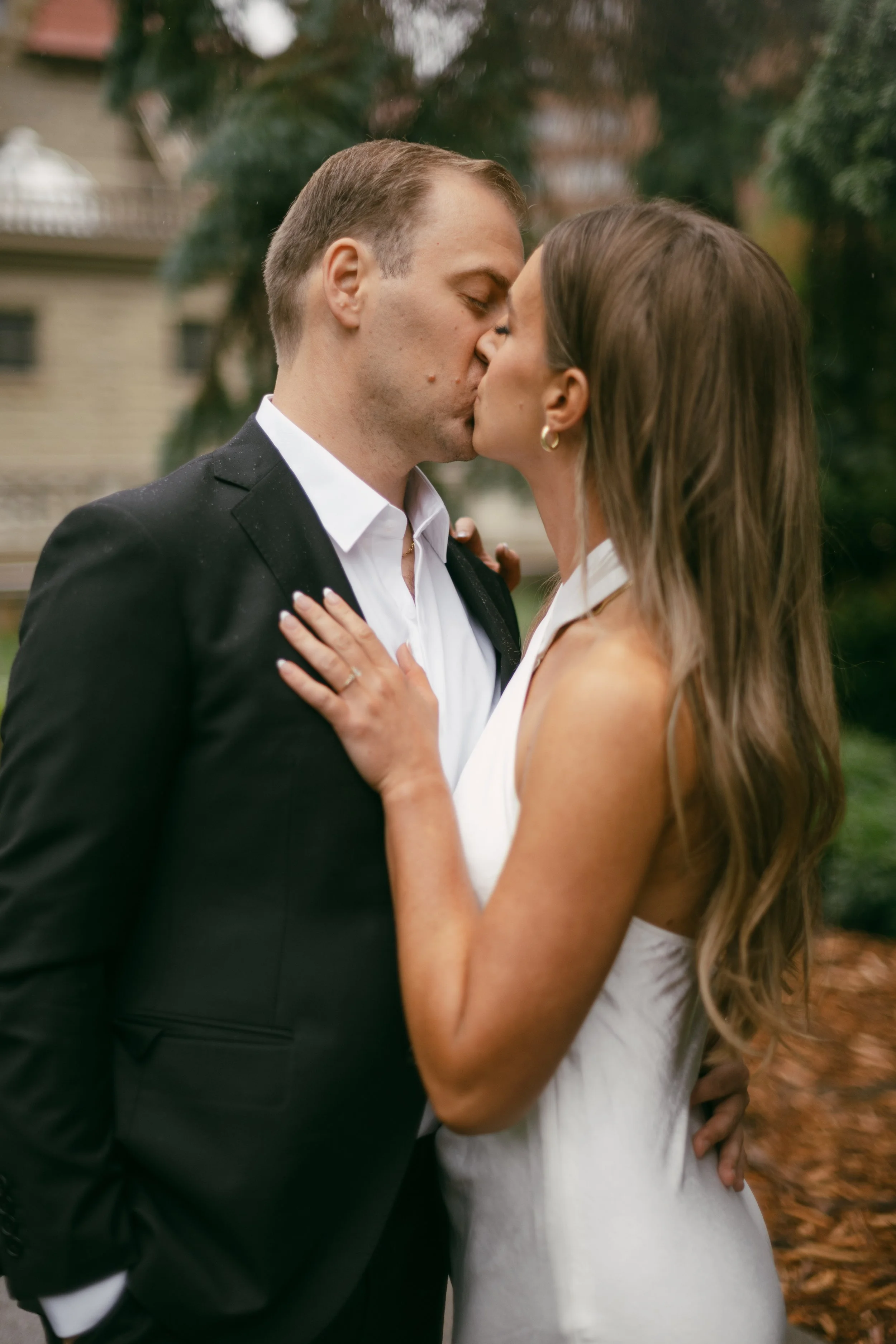 A couple sharing a kiss outdoors, dressed in formal attire, with trees and a building in the background.