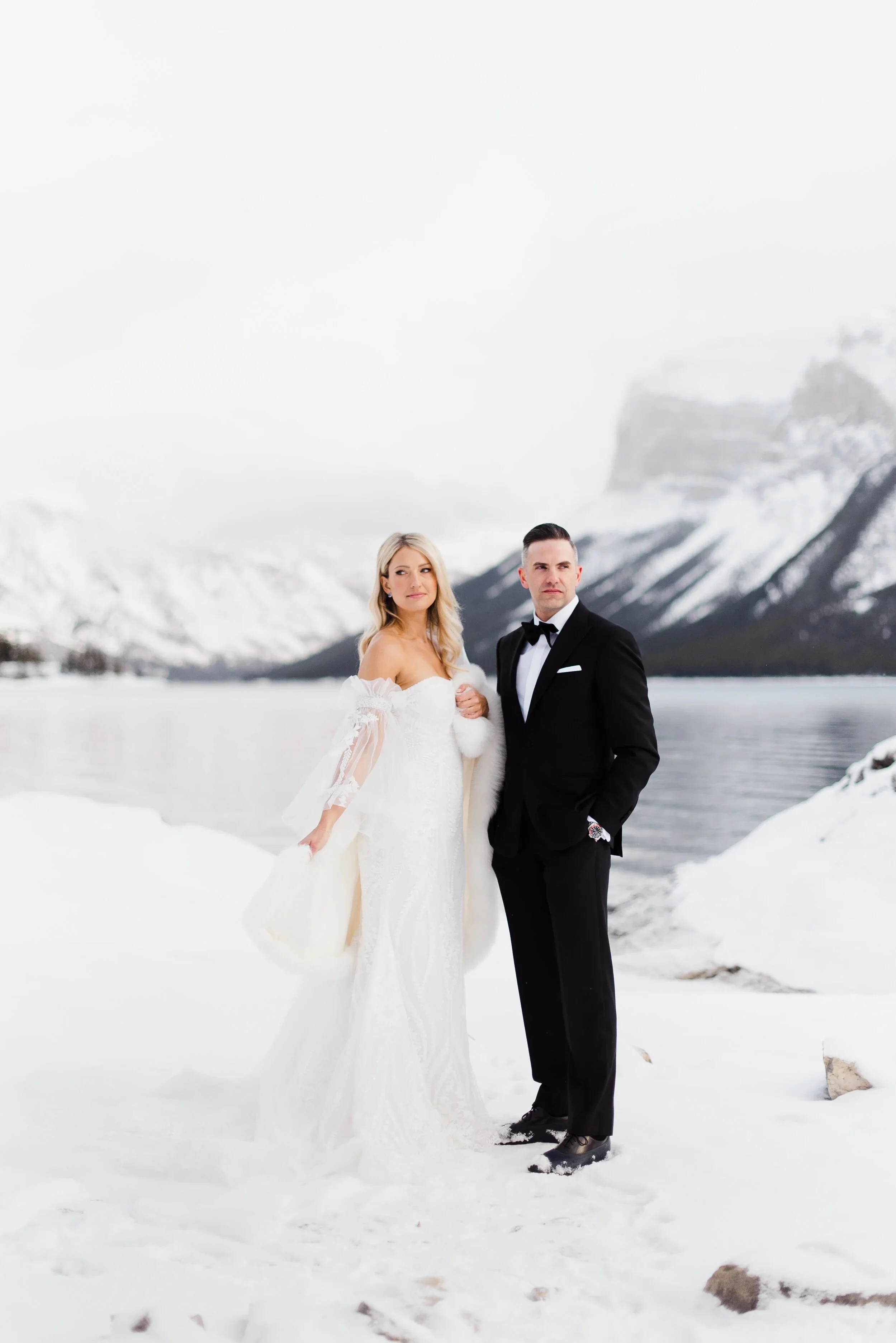 Bride and groom in wedding attire standing in snowy landscape with mountains and lake in background, winter setting.