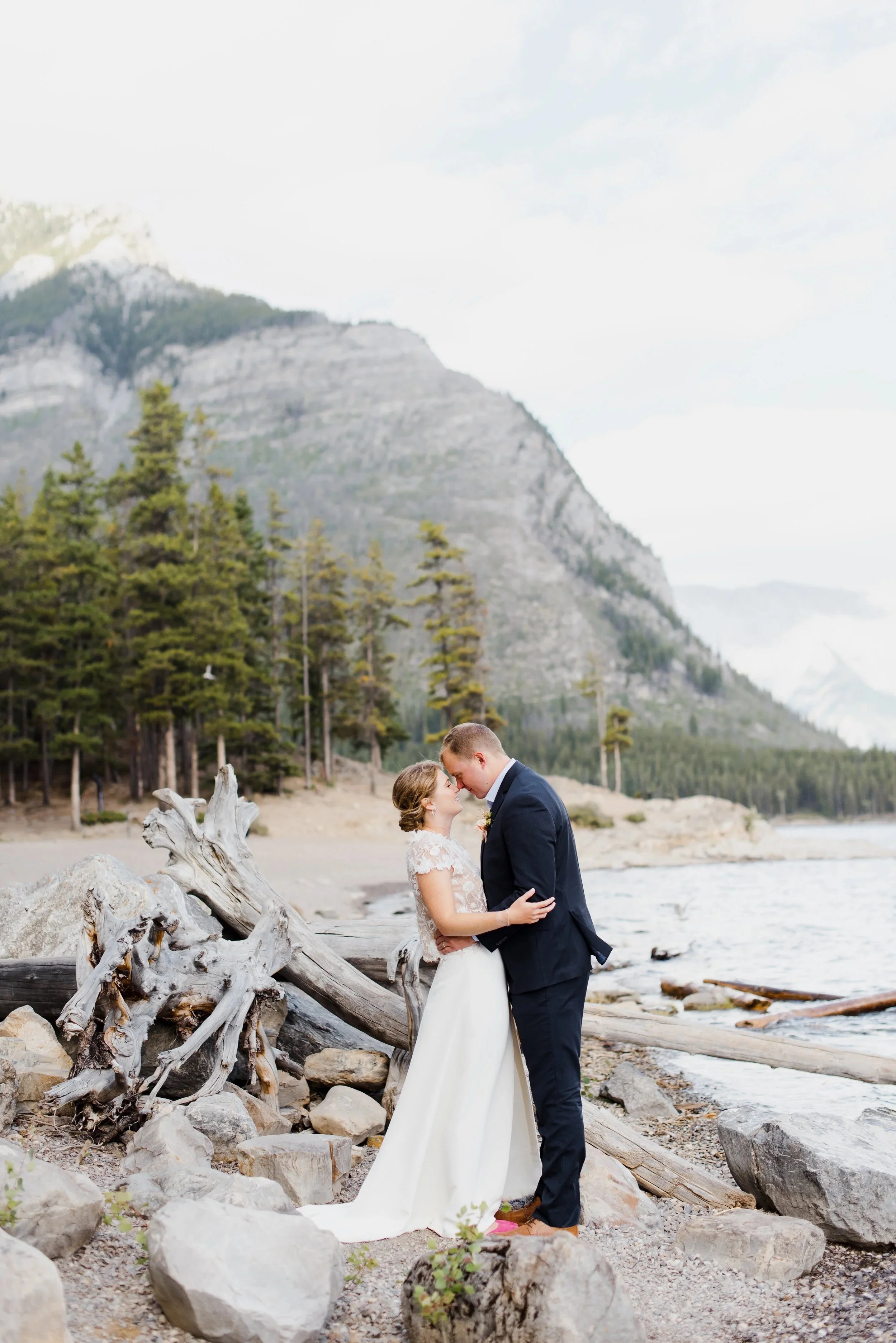 A couple in wedding attire standing on a rocky beach, embracing each other with a mountain and forest in the background.