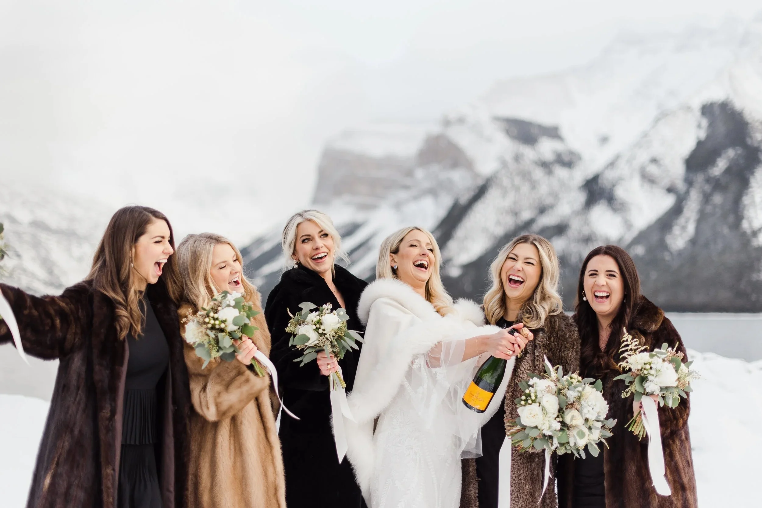 A wedding celebration outdoors in a snowy mountain landscape, featuring a bride in a white dress and fur-lined cape holding a bottle of champagne, surrounded by five women in fur coats holding bouquets of flowers, all laughing and smiling.