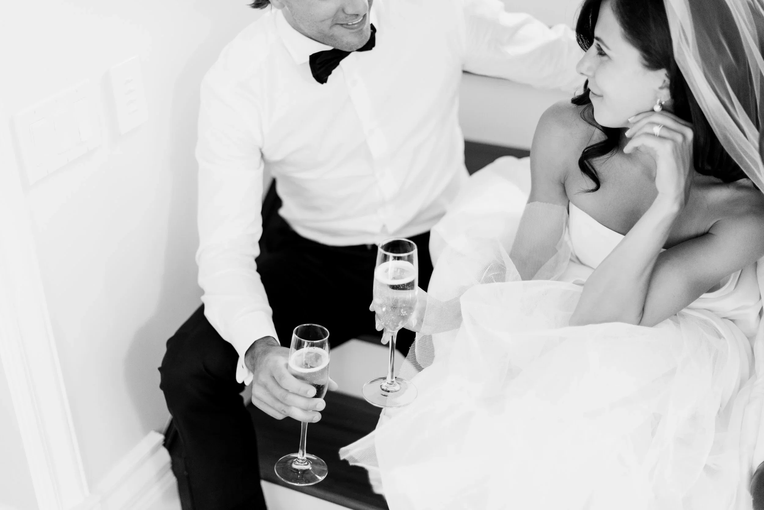 A bride and groom sitting together, clinking glasses of champagne, smiling at each other.