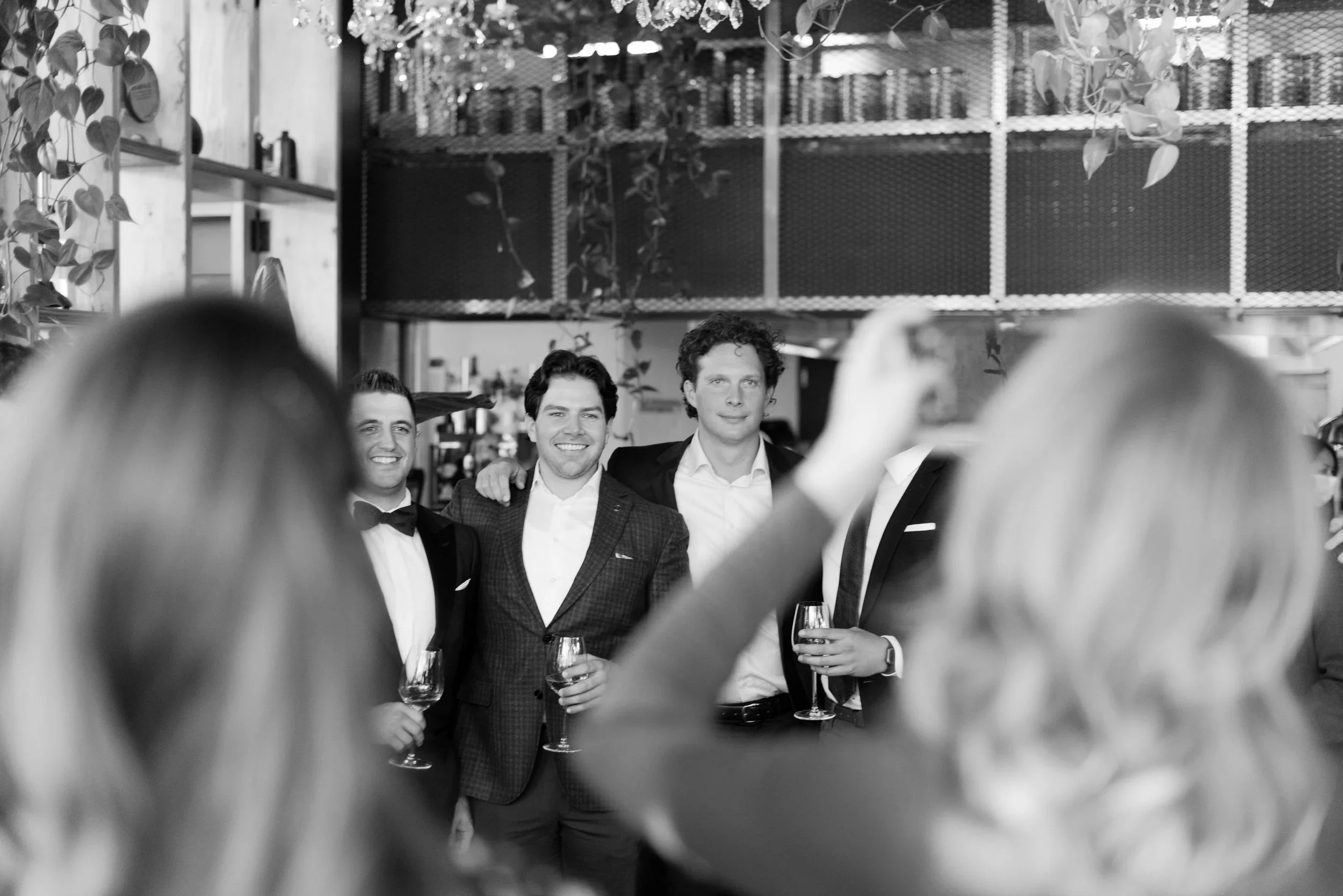 Black and white photo of four men in formal suits posing for a picture, with two women in the foreground taking the photo at what appears to be a celebration or event.