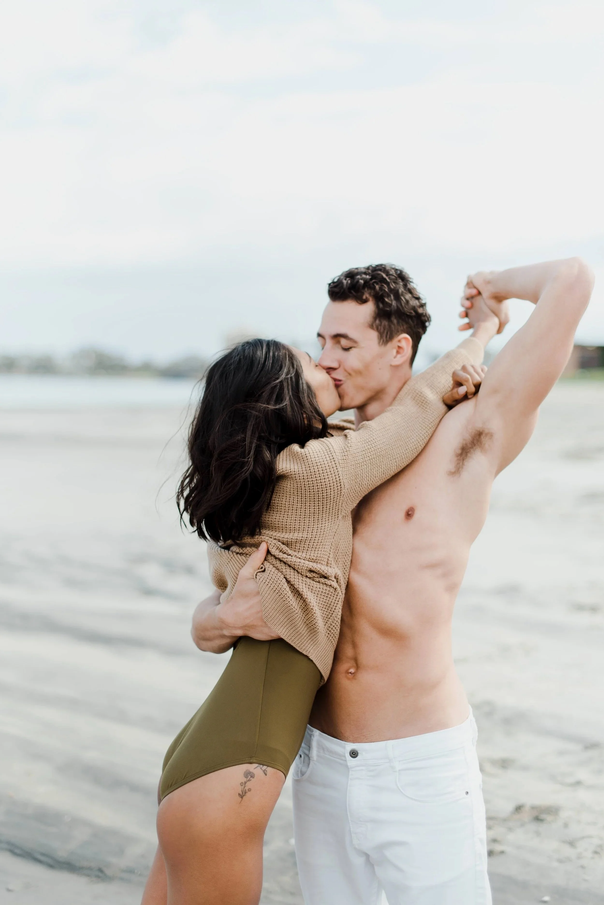 A young couple sharing a kiss on a beach, with the woman in shorts and a sweater and the man shirtless, holding each other closely.