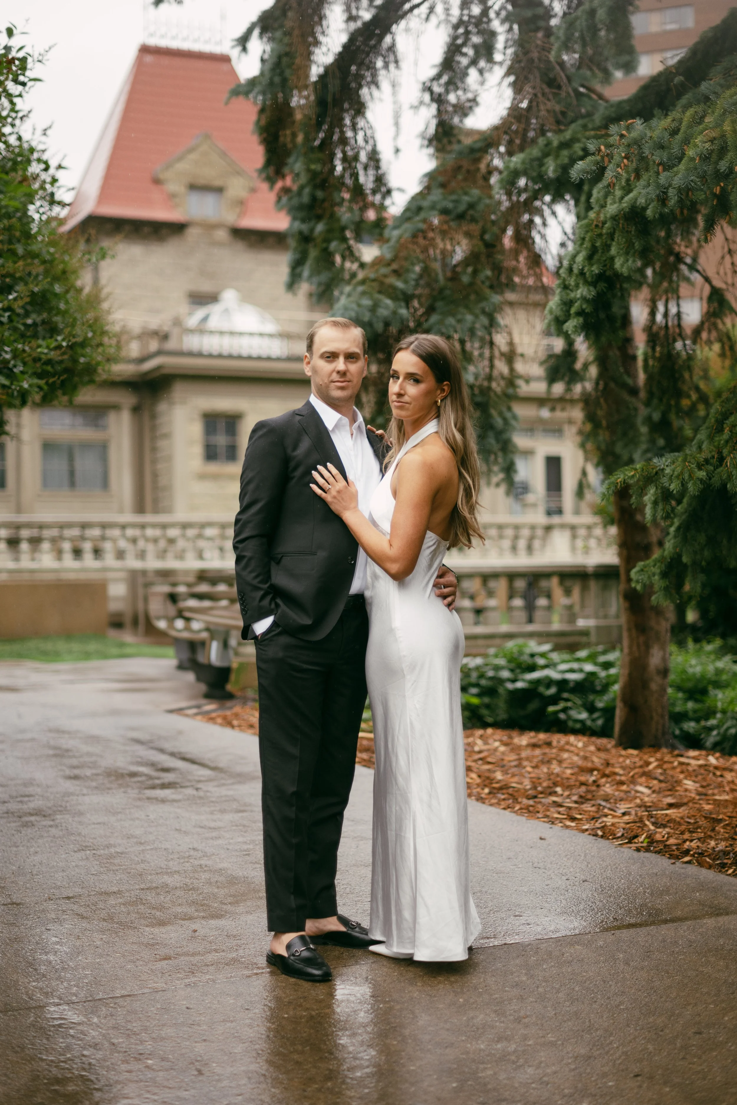 A couple dressed in formal wedding attire standing outdoors on a wet pathway with a historic building and trees in the background.