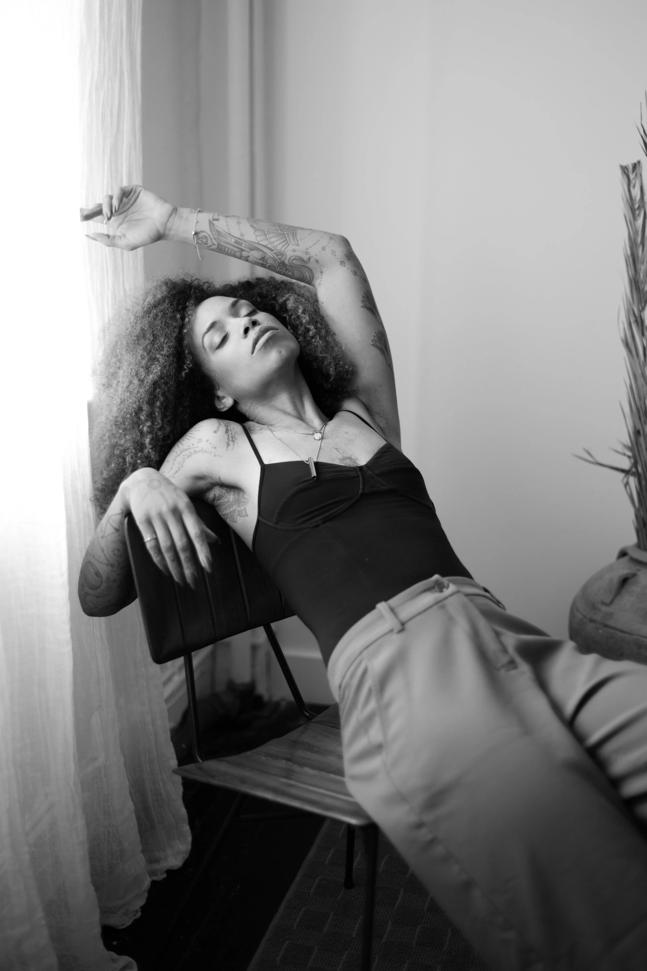A woman with curly hair and tattoos, wearing a black top and beige pants, is reclining on a chair, with her eyes closed and one arm raised above her head, in a relaxed pose.