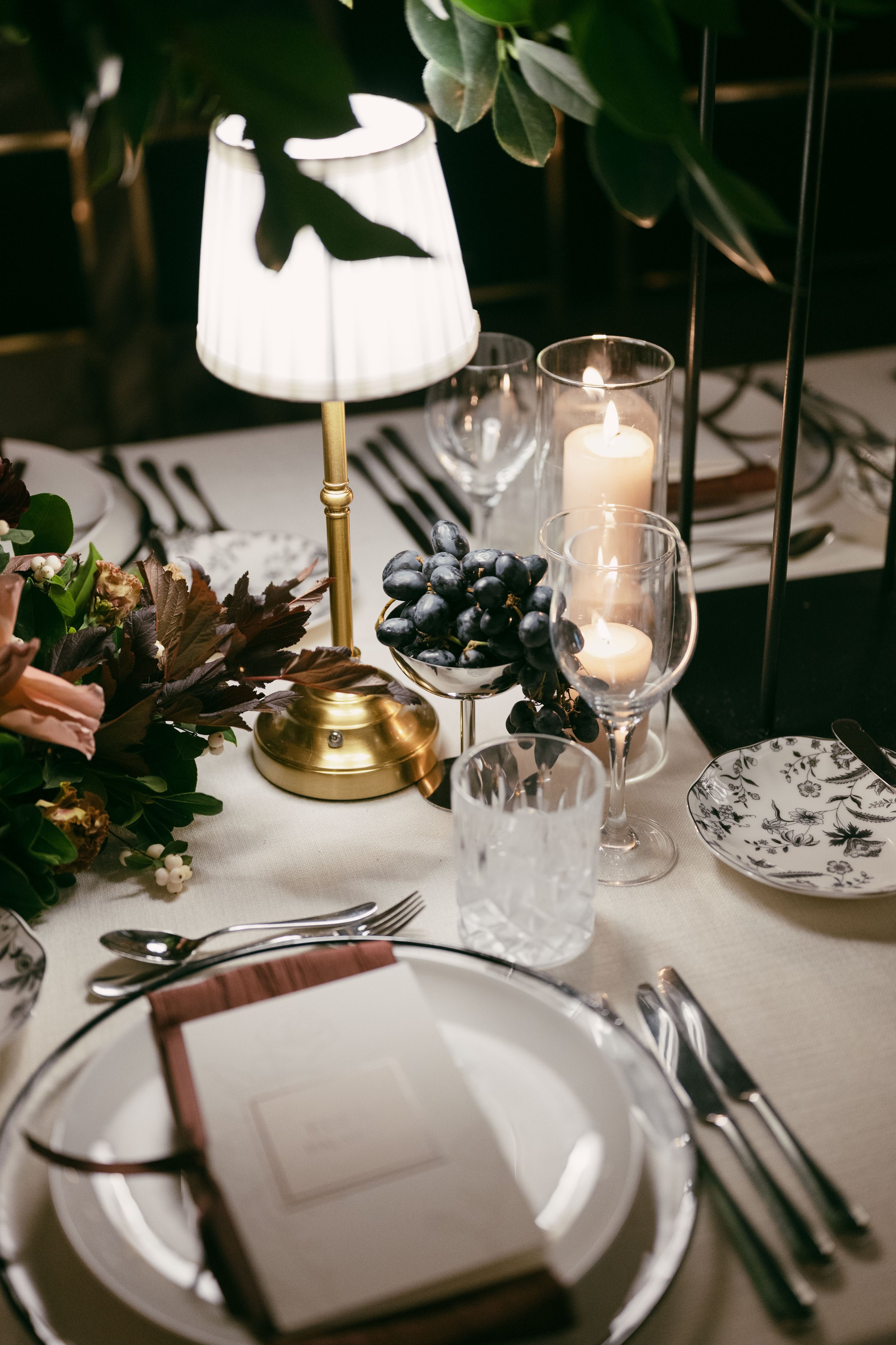 Elegant table setting with a gold lamp, candles, grapes, floral arrangement, and fine dinnerware.