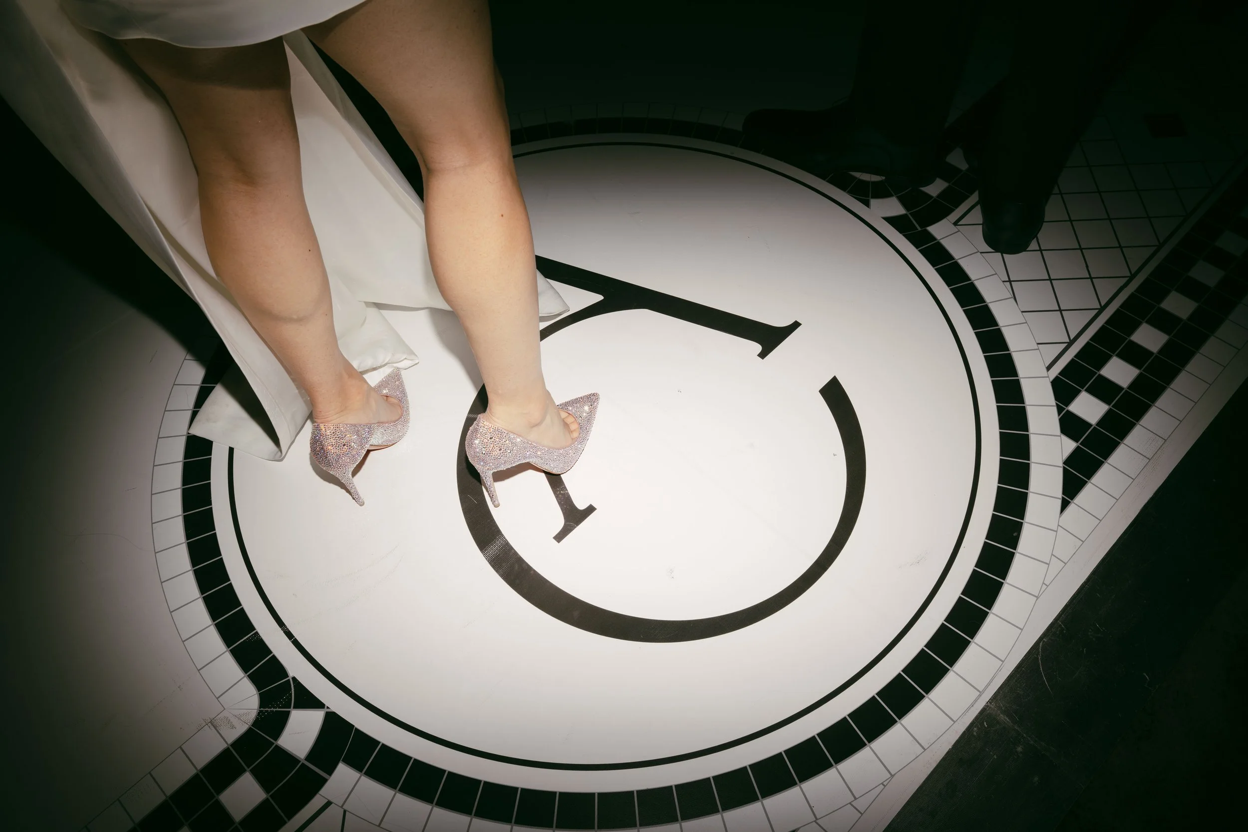 A woman in high-heeled shoes standing on a large clock face with black and white tiles, viewed from above.
