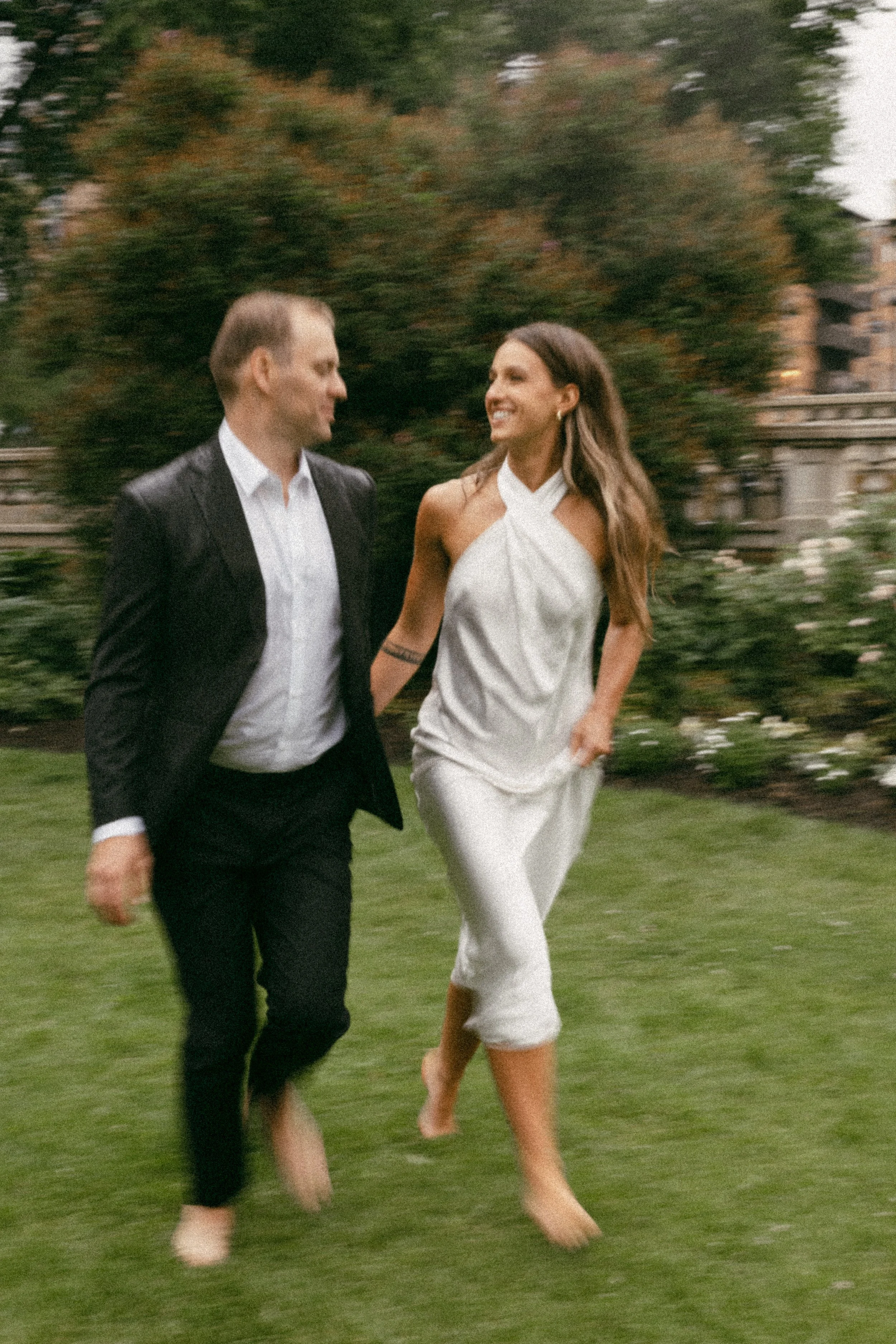 A man and woman walking barefoot on a lawn, smiling and looking at each other, surrounded by greenery and trees in a garden or park setting.