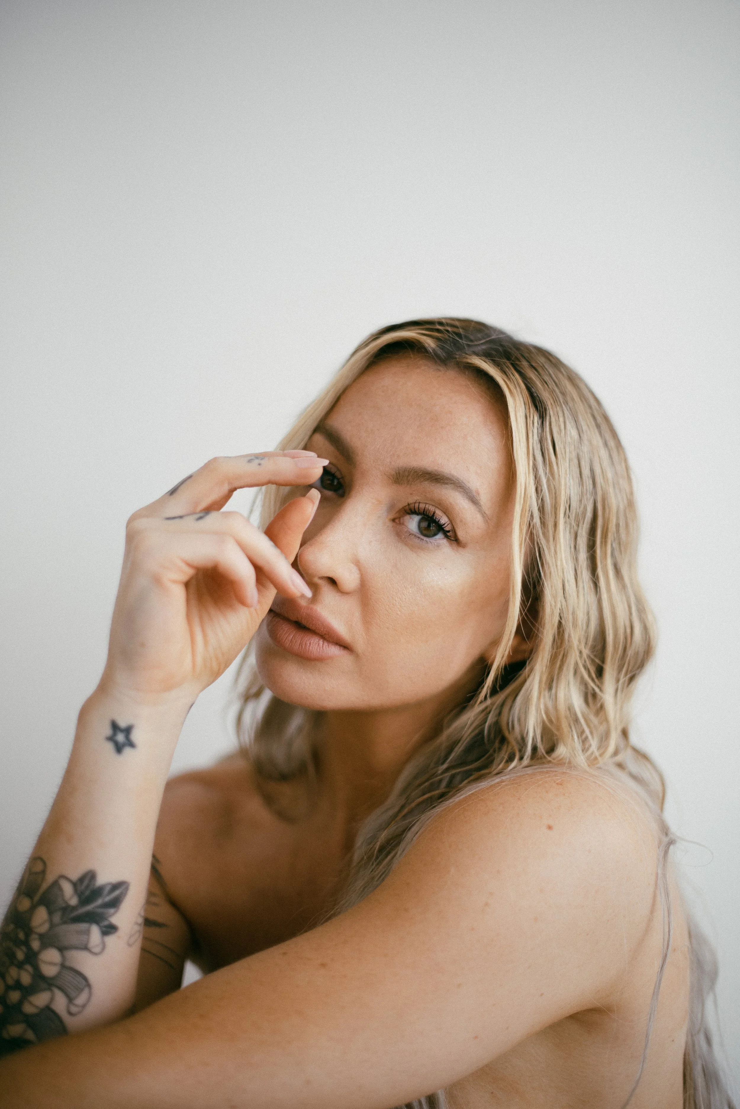 A woman with long wavy blonde hair and tattoos on her arm, looking at the camera with her hand near her face, in front of a plain white background.