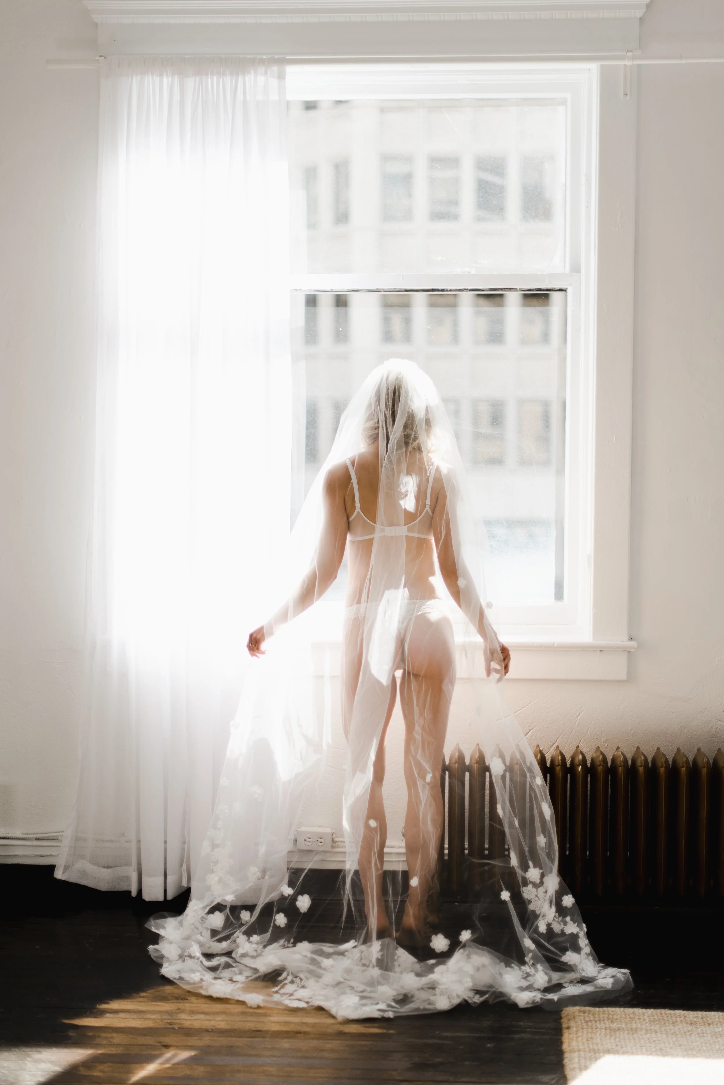 Woman in a wedding veil and lingerie standing by a window with sunlight streaming in, white curtains, wooden floor, radiator, and light-colored wall.