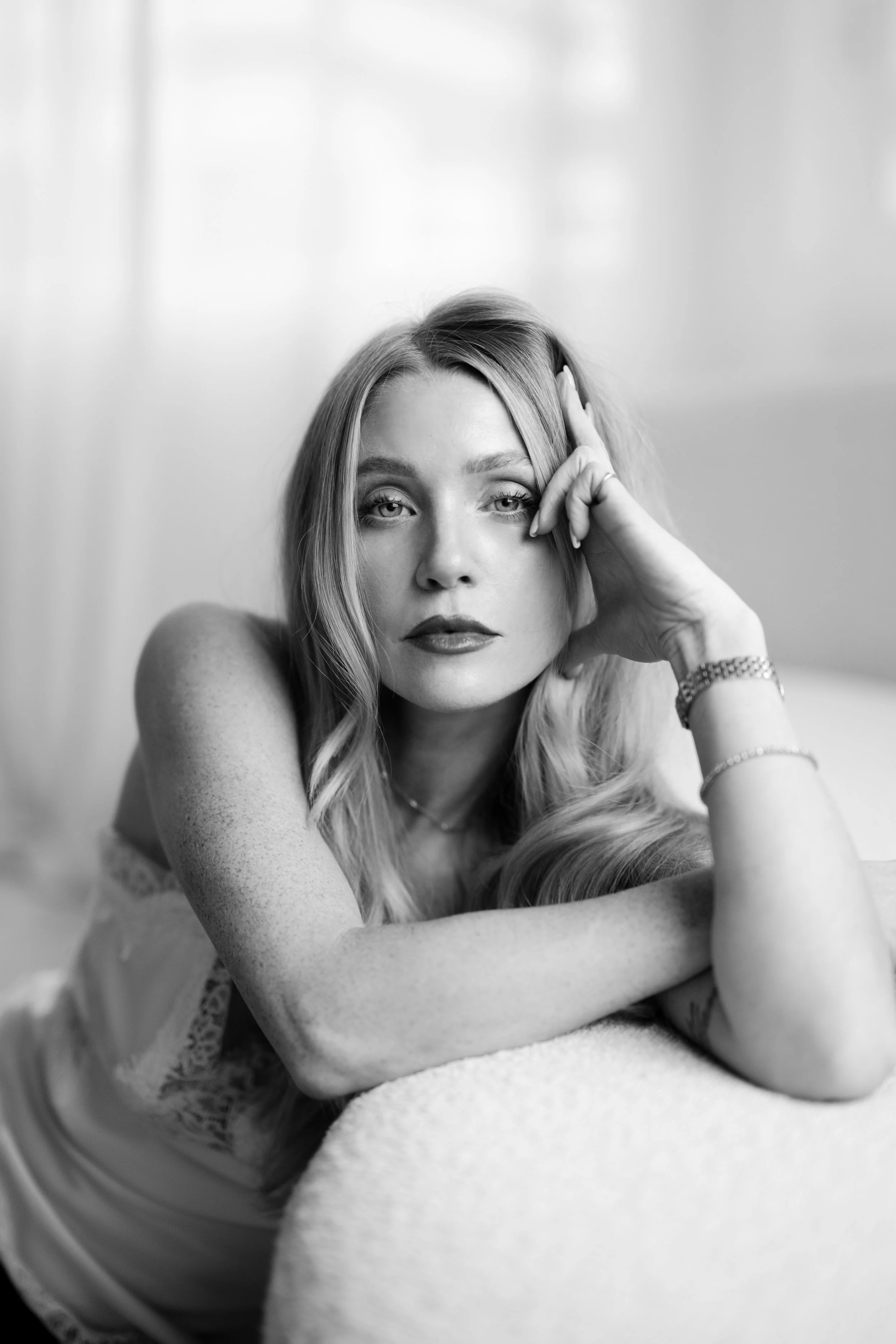 Black and white portrait of a young woman with long, wavy hair, resting her head on her arm, looking directly at the camera with a relaxed expression.