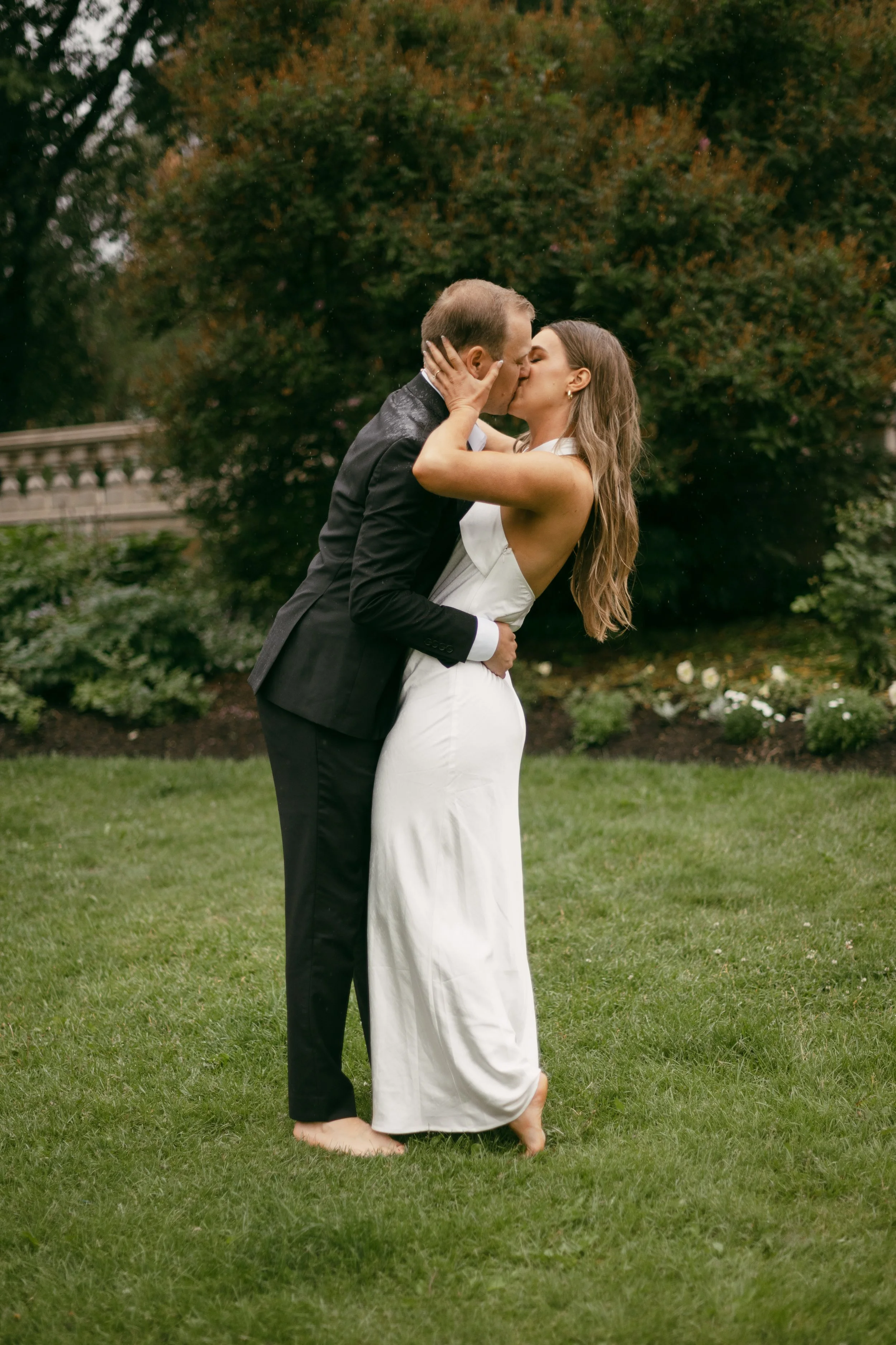A couple sharing a kiss outdoors on a grassy lawn, with the man wearing a black suit and the woman in a white dress.