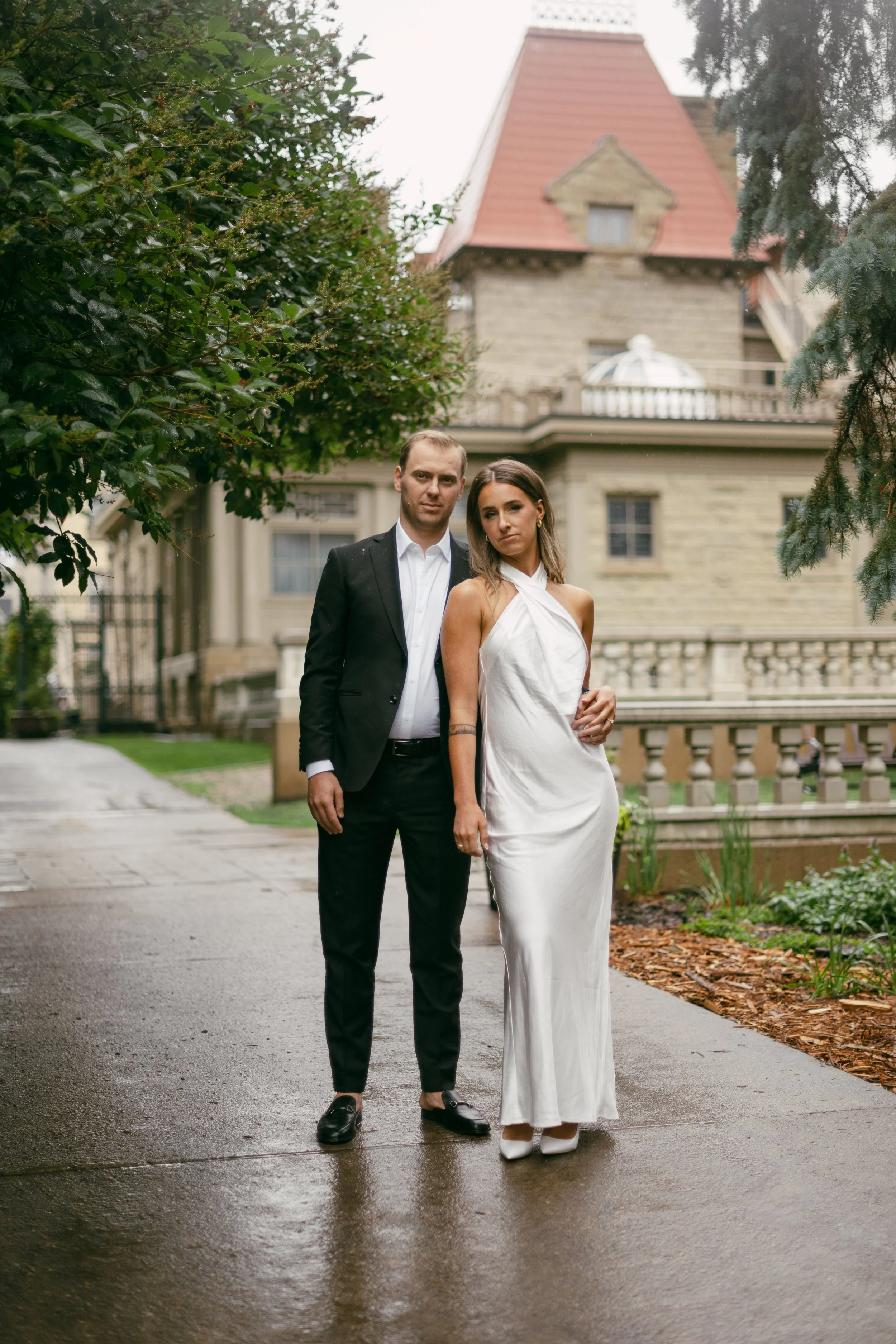 A man and woman are standing outdoors on a wet sidewalk in front of a large house with a turret and a wraparound porch. The man is in a black suit and white shirt, and the woman is wearing a long white dress. Both are looking at the camera.