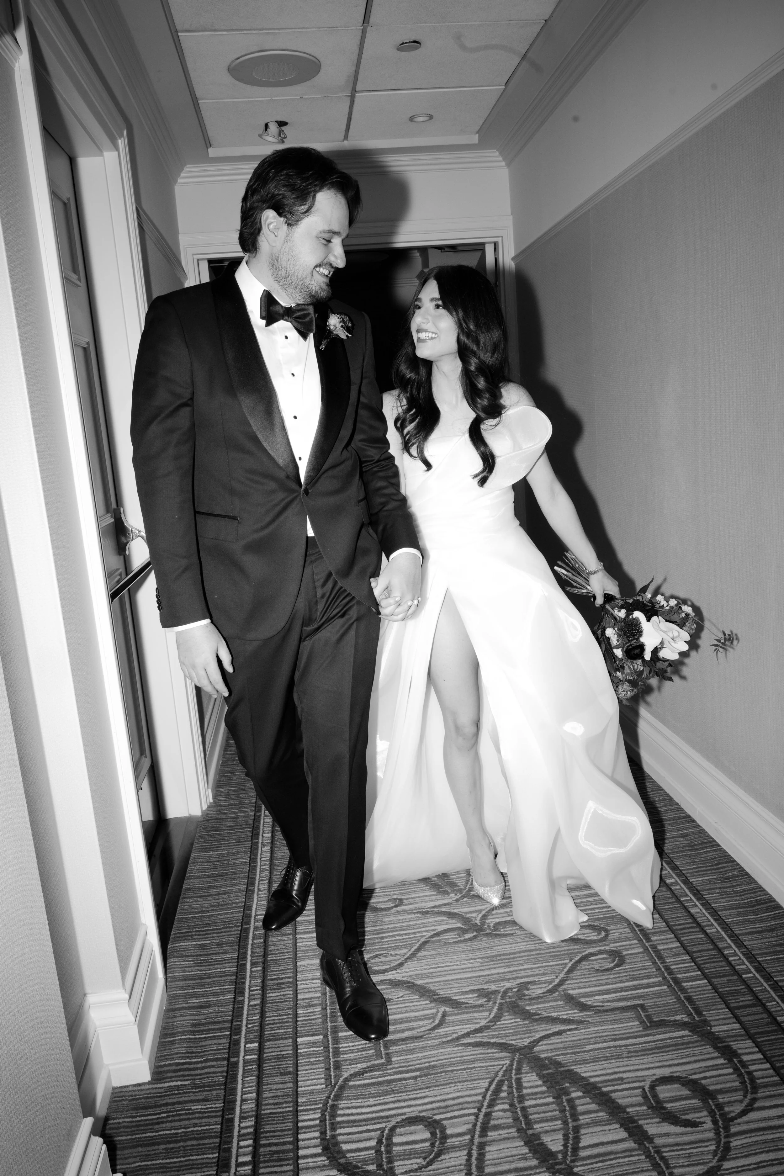A newlywed couple holding hands and smiling in a hallway, with the bride holding a bouquet of flowers, both dressed in wedding attire.