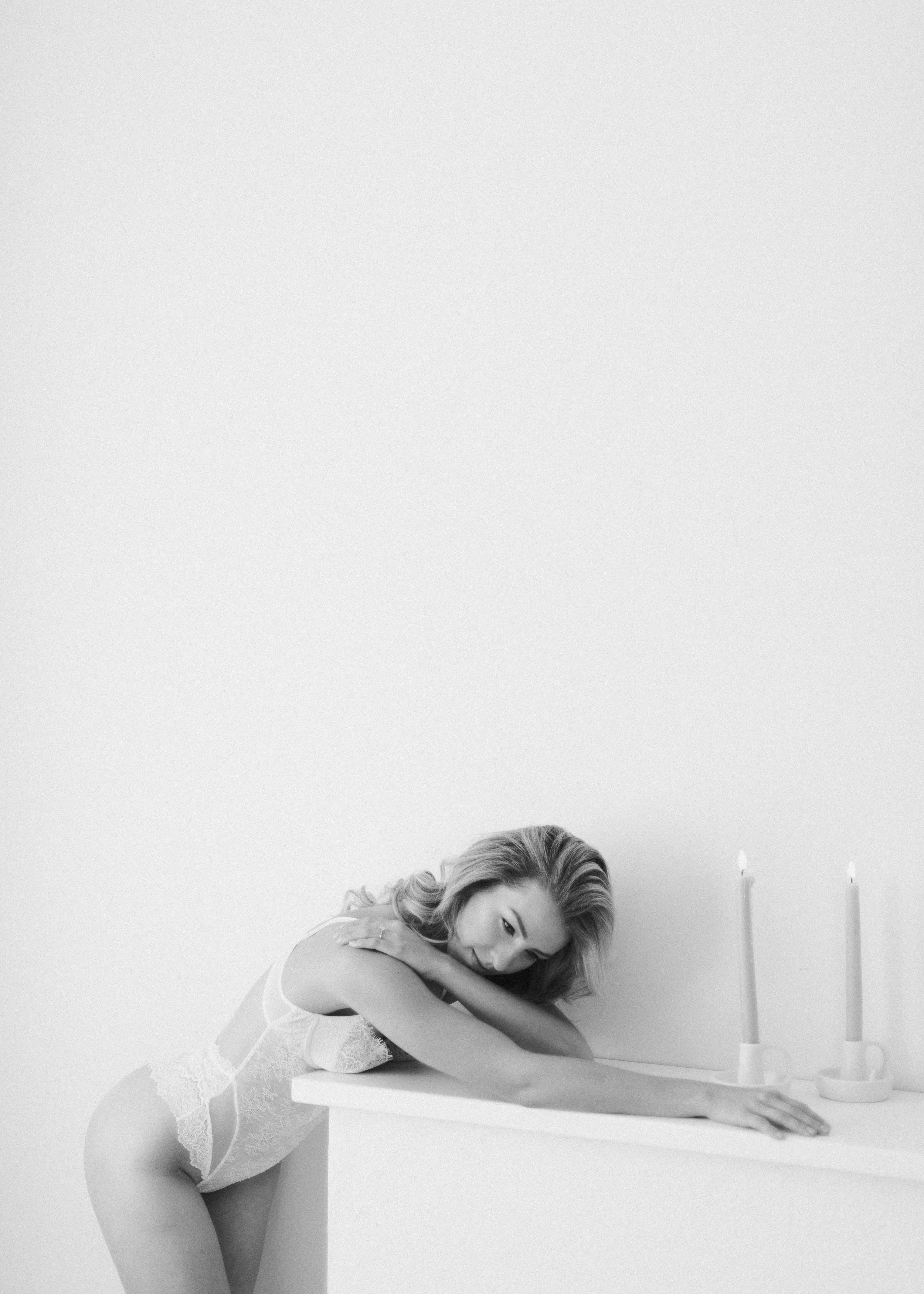 A woman in lingerie leaning on a white surface with three lit candles behind her in a minimalistic setting.