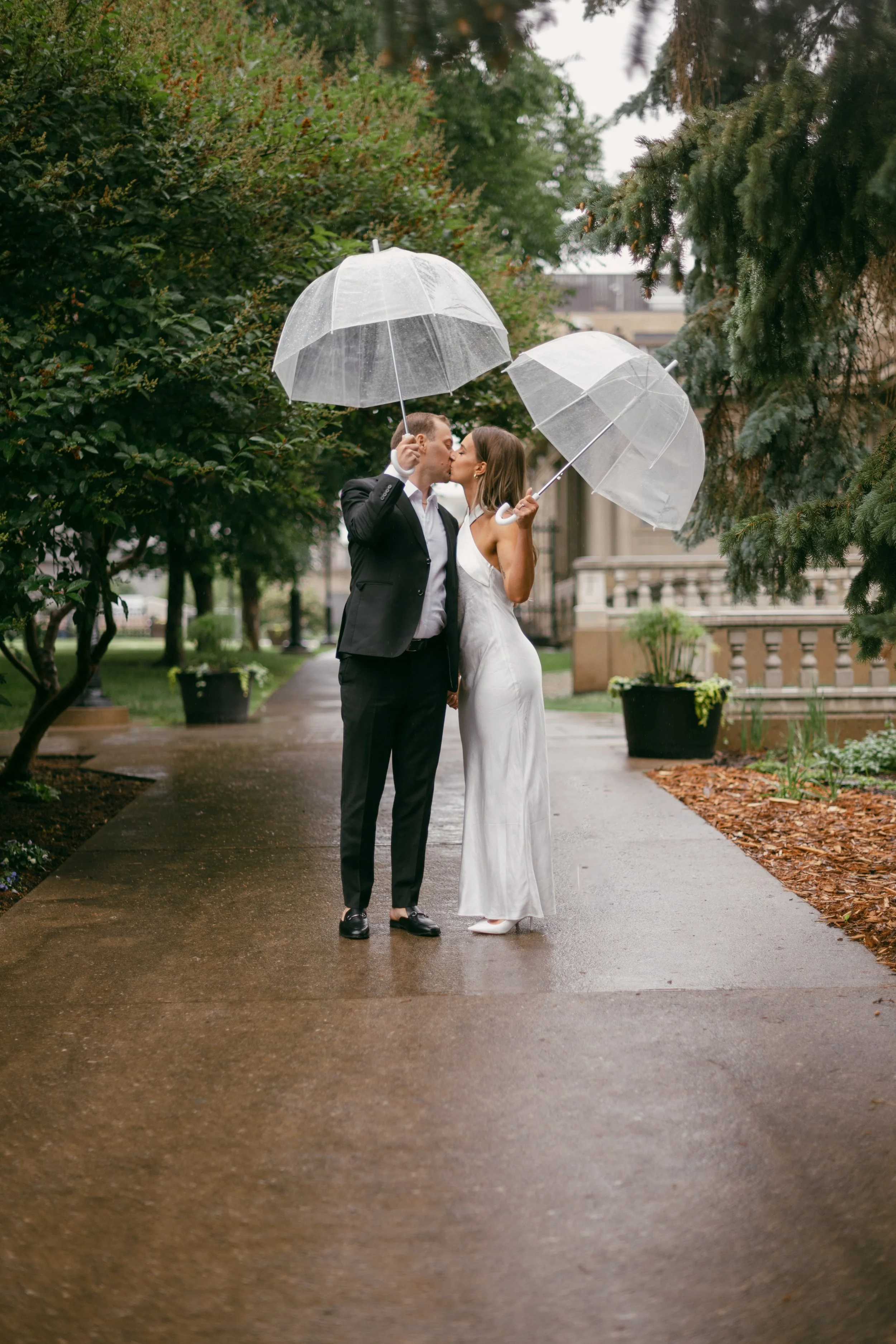A couple dressed in wedding attire kissing under clear umbrellas on a rainy day in a garden pathway.